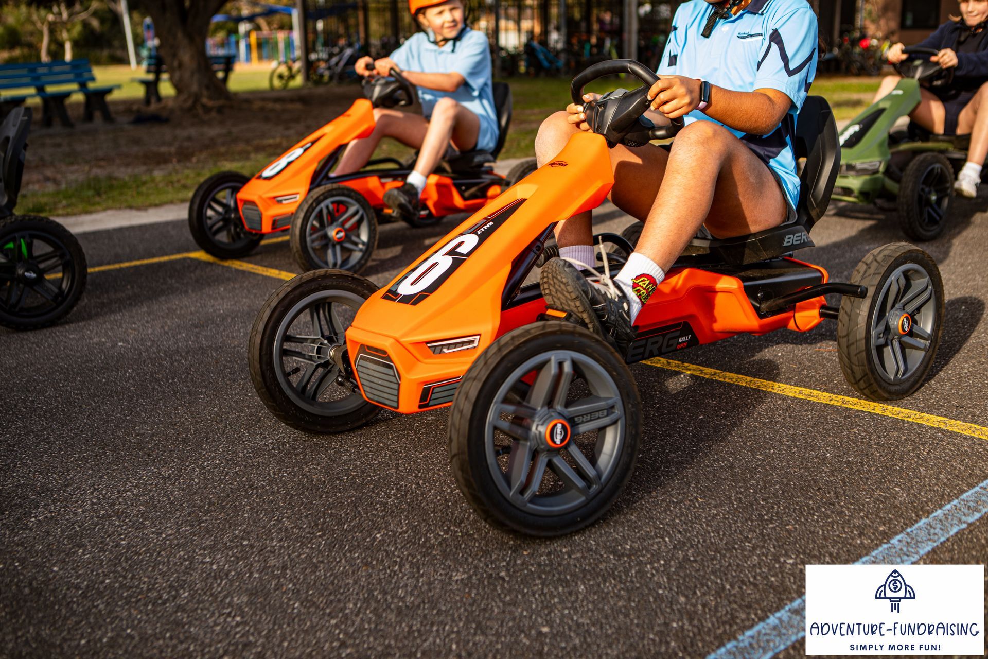 People riding orange go-karts on a paved surface, likely an outdoor school activity.