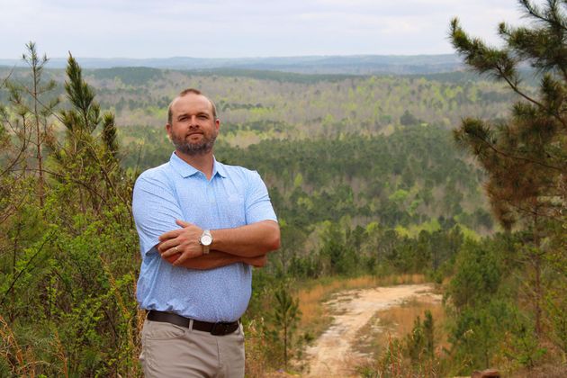 A person with arms crossed stands outdoors on a hill overlooking a vast, forested valley.