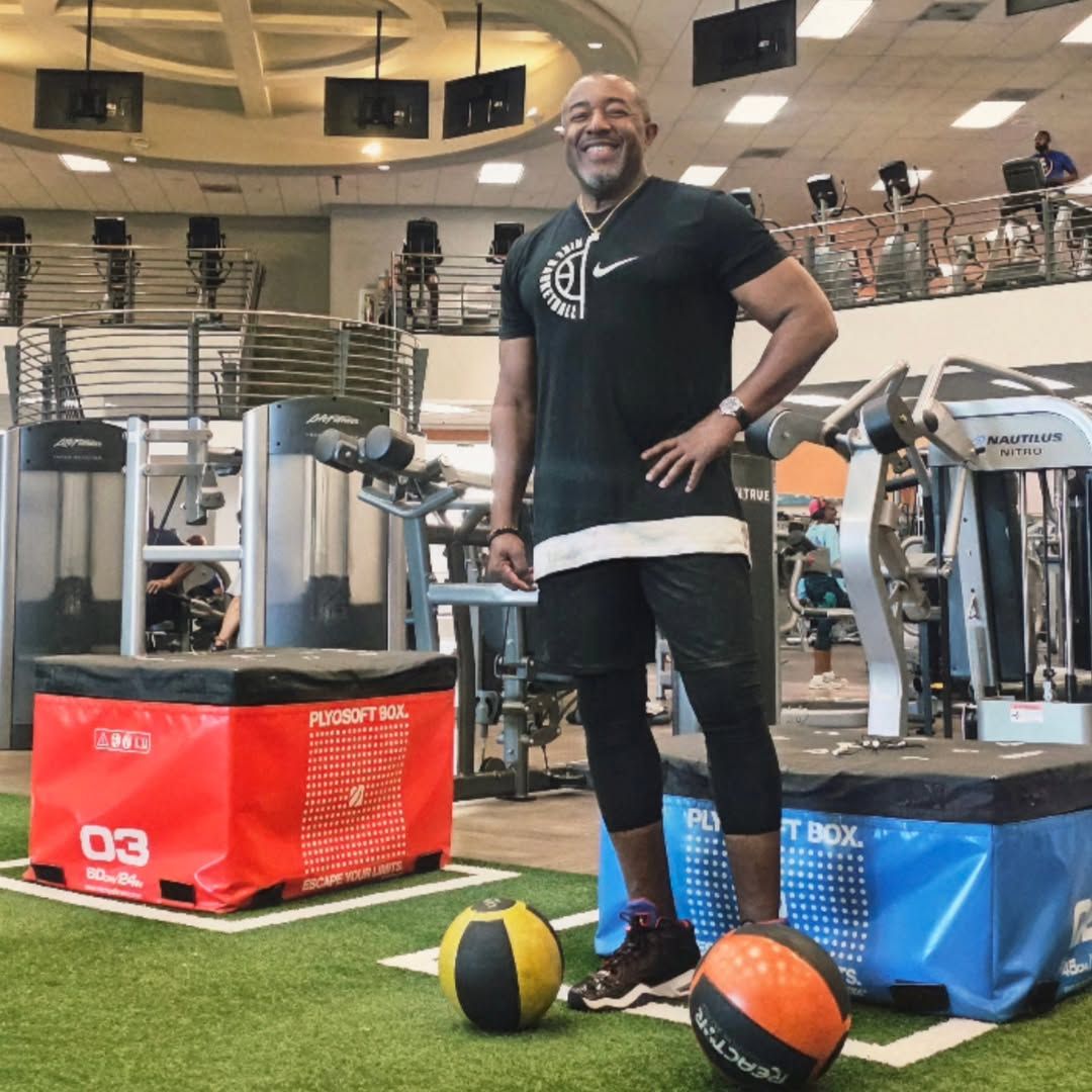 Man in gym attire poses in a gym. He's wearing black clothing, smiling, and has his hand on his hip.
