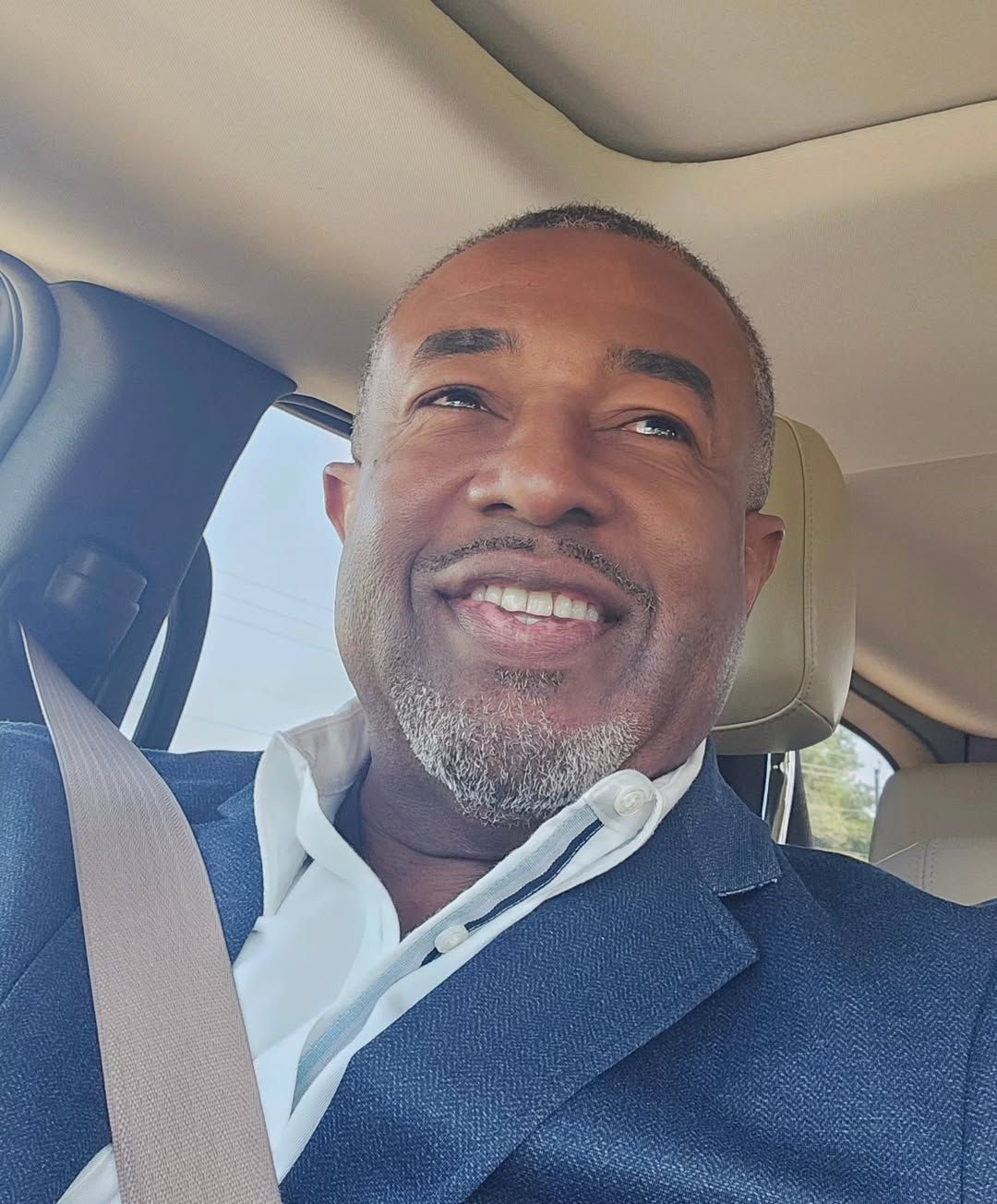 Man in a car smiling, wearing a blue blazer and seatbelt, looking upward.
