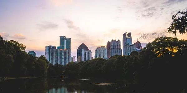 City skyline at sunset seen across a dark lake, trees frame the bottom.