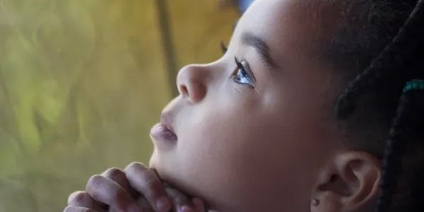 A young child with folded hands looks up, eyes focused. Their expression is thoughtful against a blurry background.