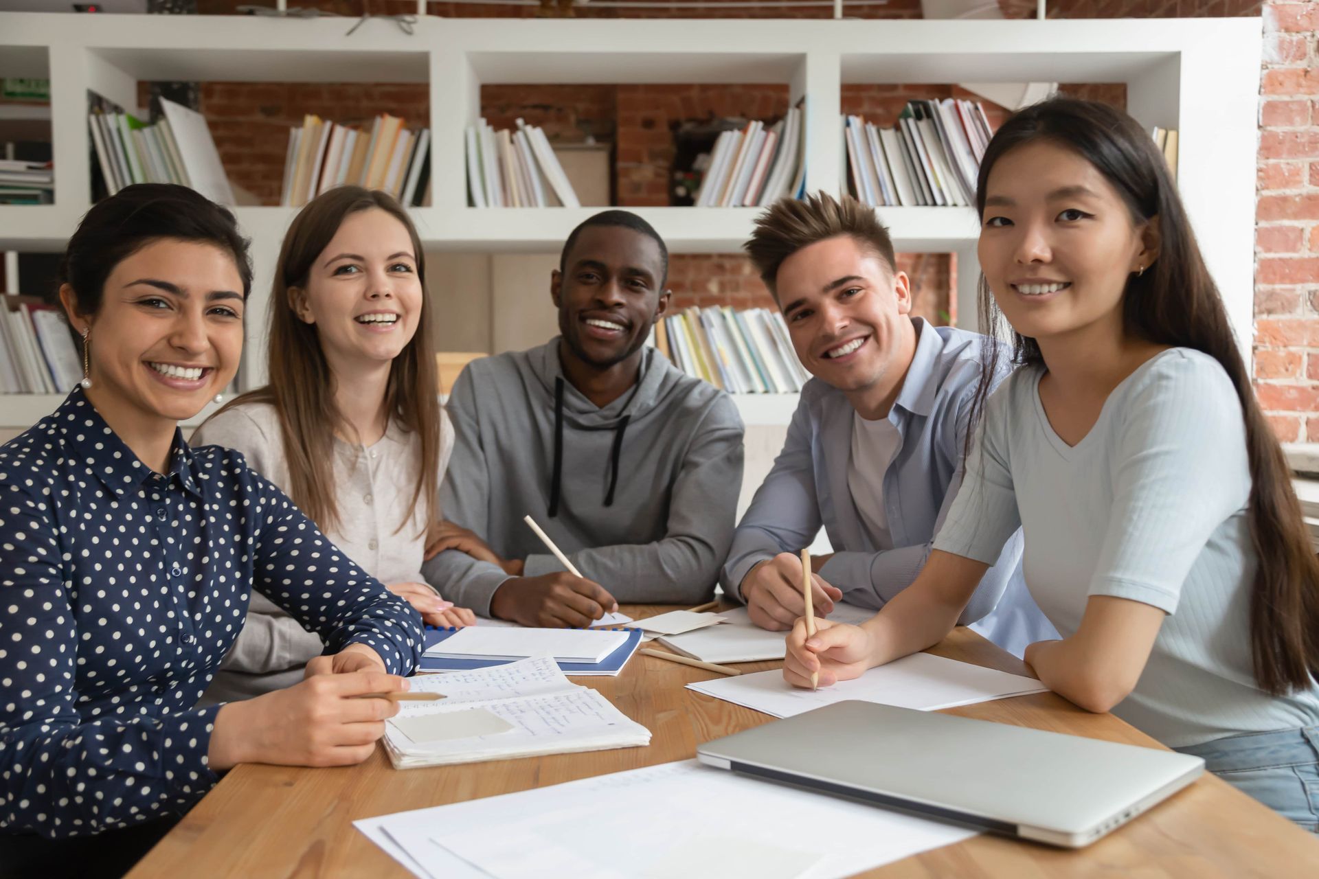 Five diverse students smiling at a table with books, papers, and a laptop in a library setting.