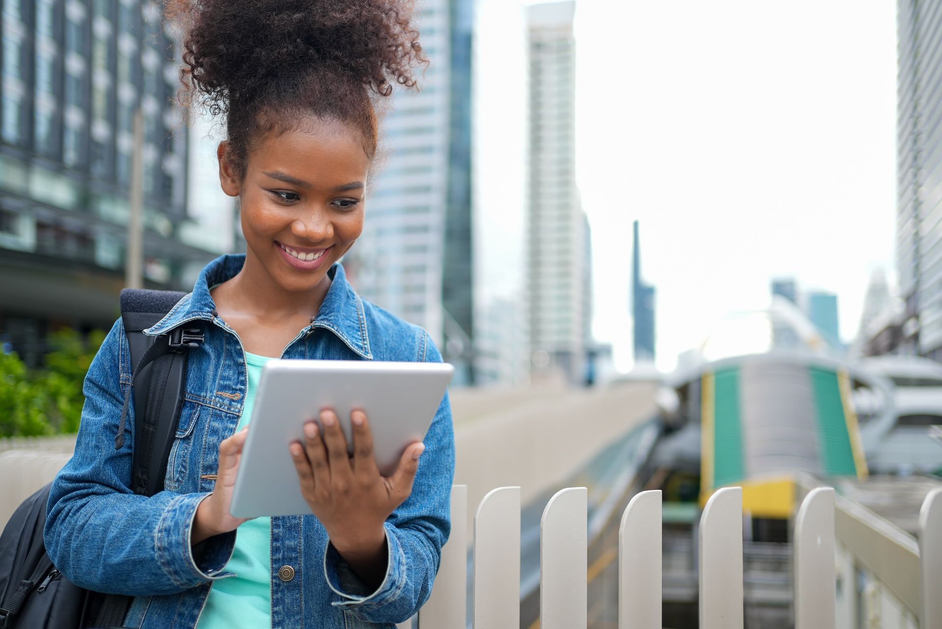 Young Black woman smiles, using a tablet outdoors.
