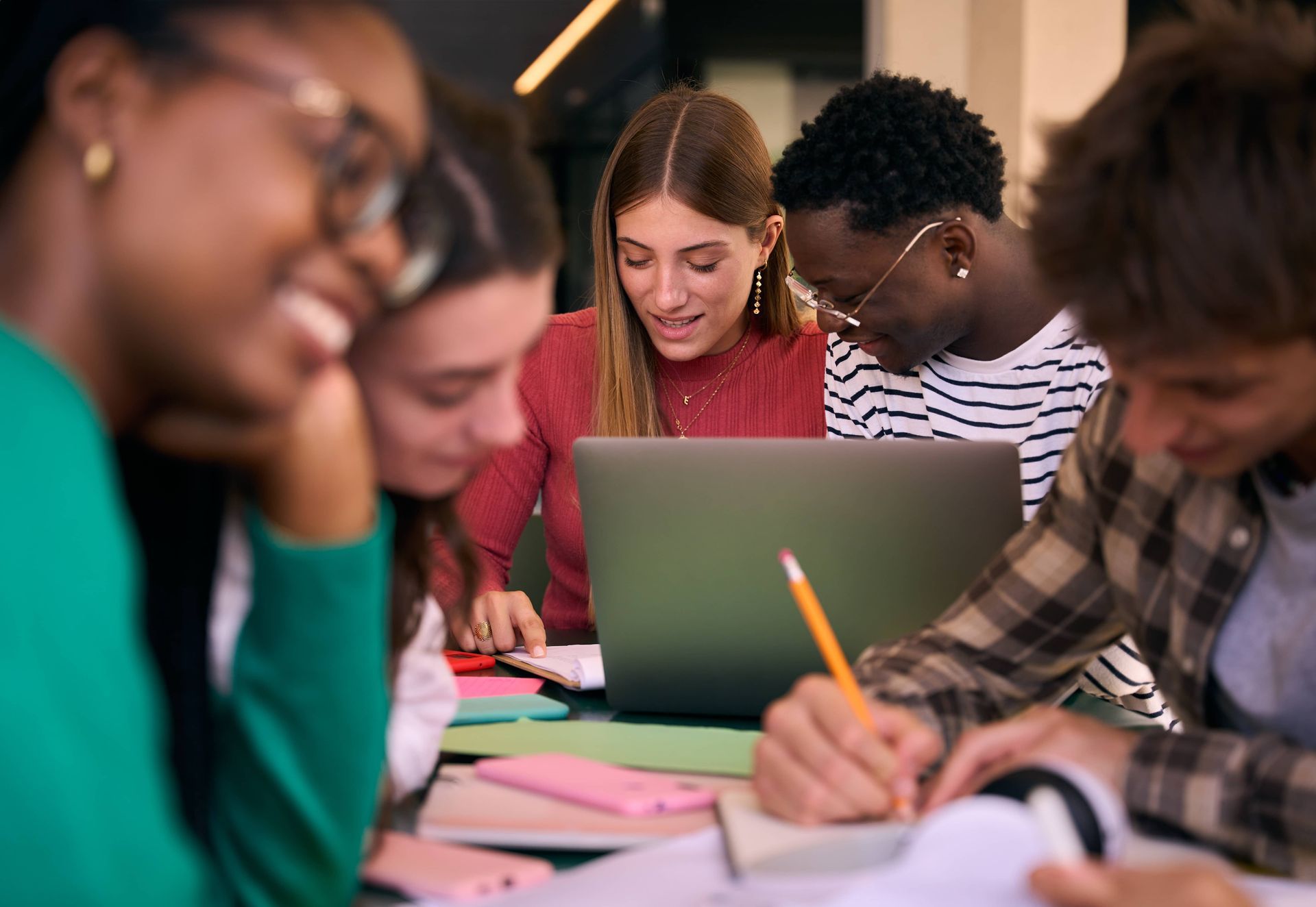Group of diverse students studying together, looking at a laptop and taking notes in a bright setting.