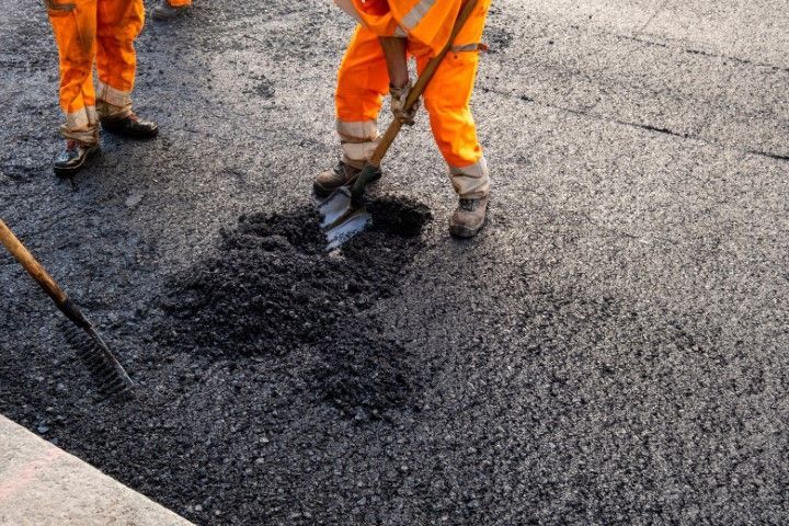 Road workers in orange safety gear spreading asphalt with shovels.