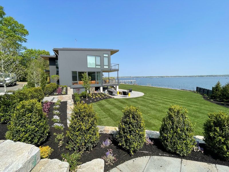 Modern gray house on a waterfront lawn; landscaped beds in foreground.