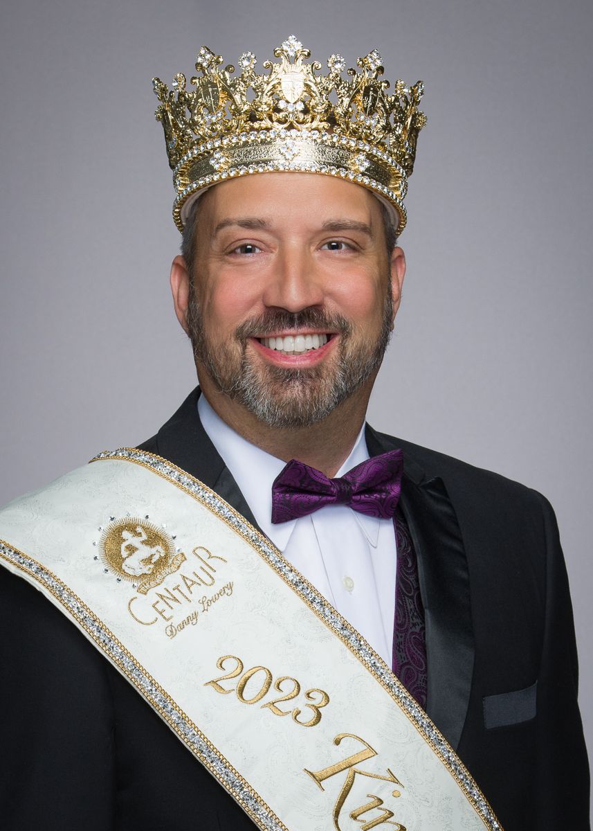 A man in a suit and bow tie is wearing a crown and sash.