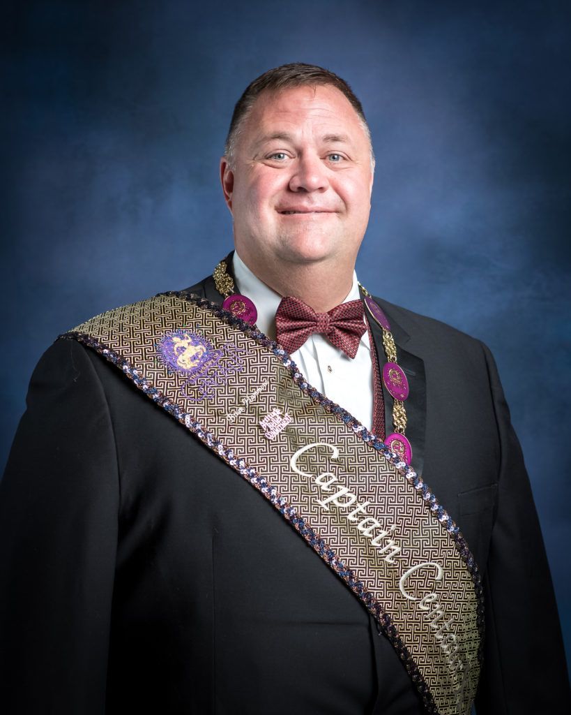 A man in a suit and bow tie is wearing a sash that says captain.