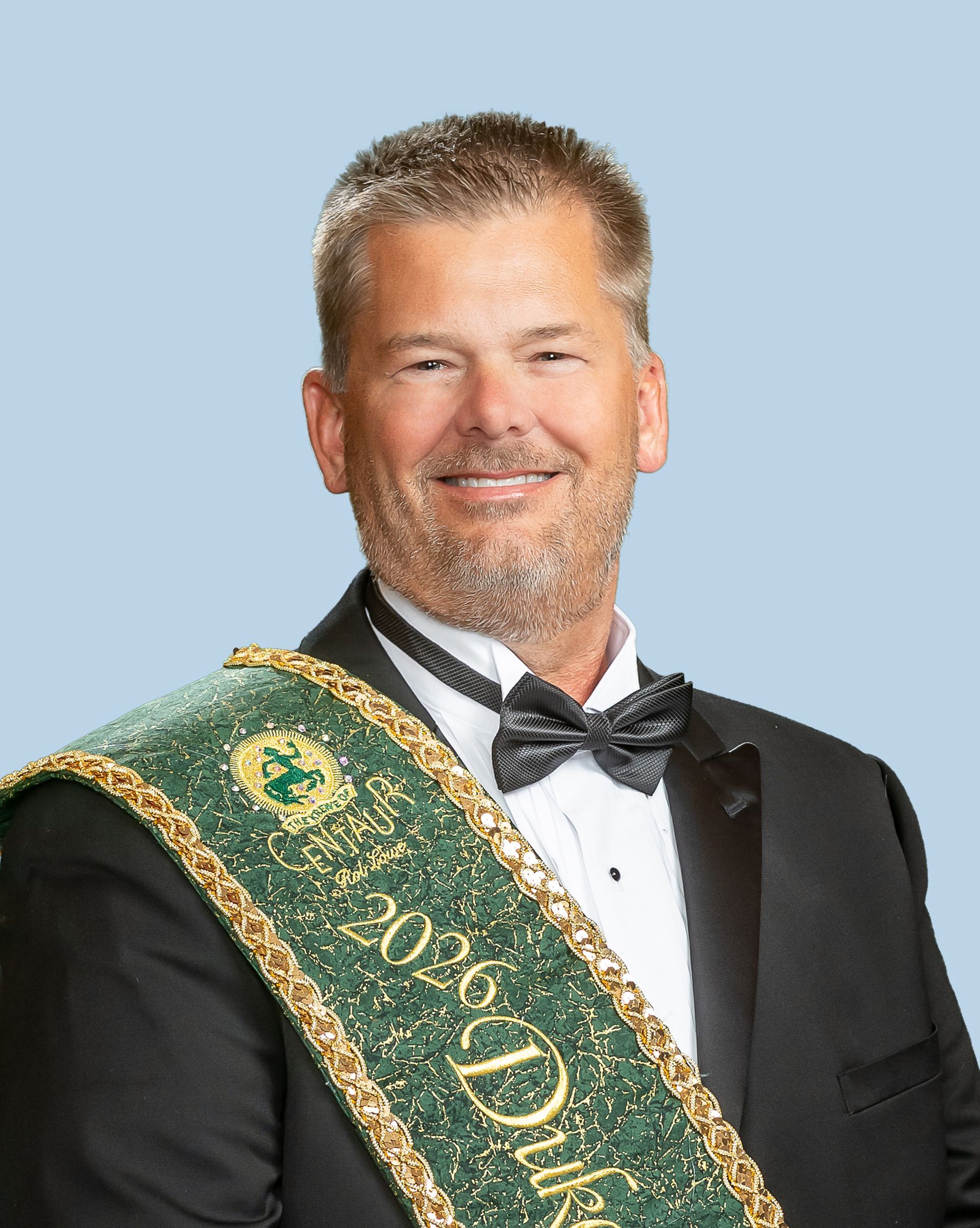 A man in a suit and bow tie is wearing a green sash.