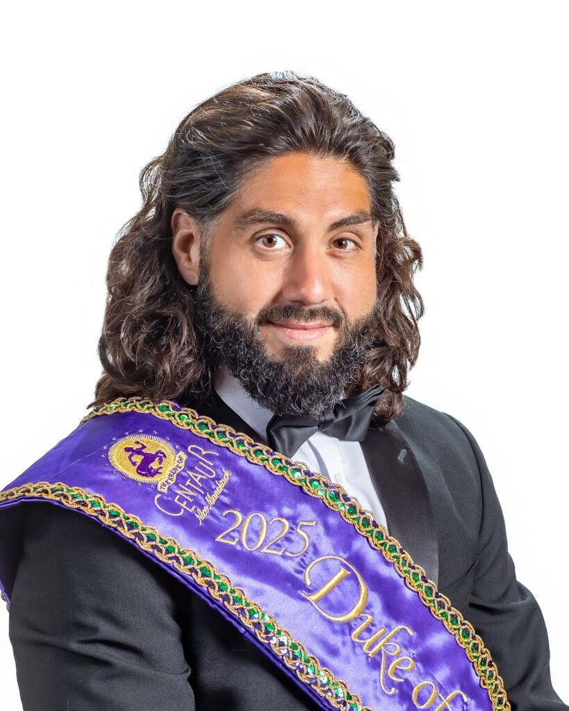 A man with long hair and a beard is wearing a sash that says duke of