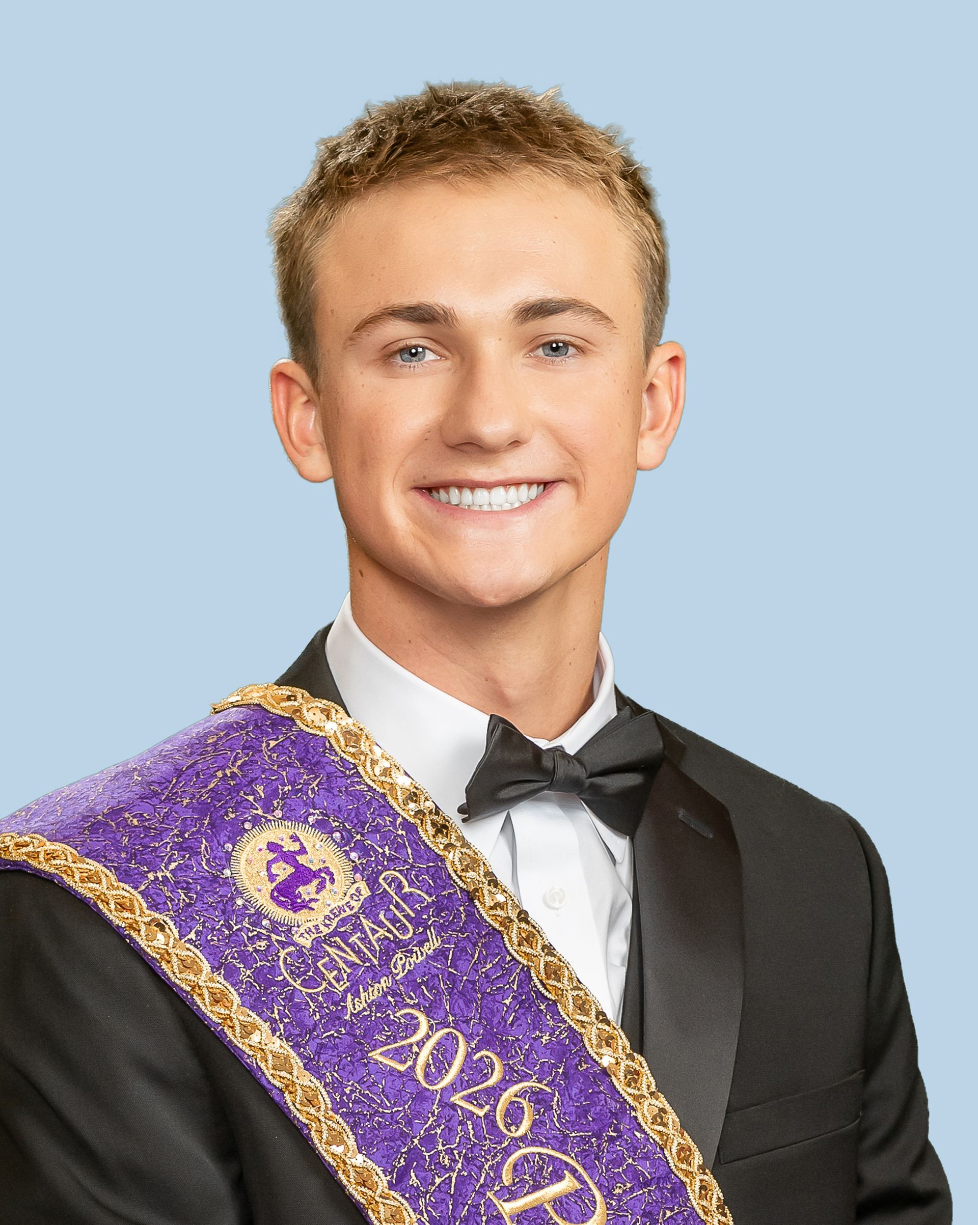 A young man in a tuxedo and bow tie is wearing a purple sash, who is a prince. 