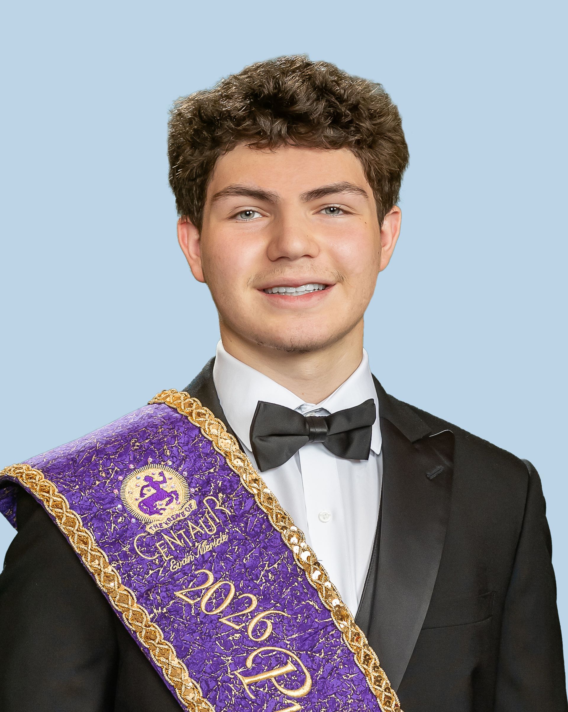 A young man in a tuxedo and bow tie is wearing a purple sash, who is a prince. 