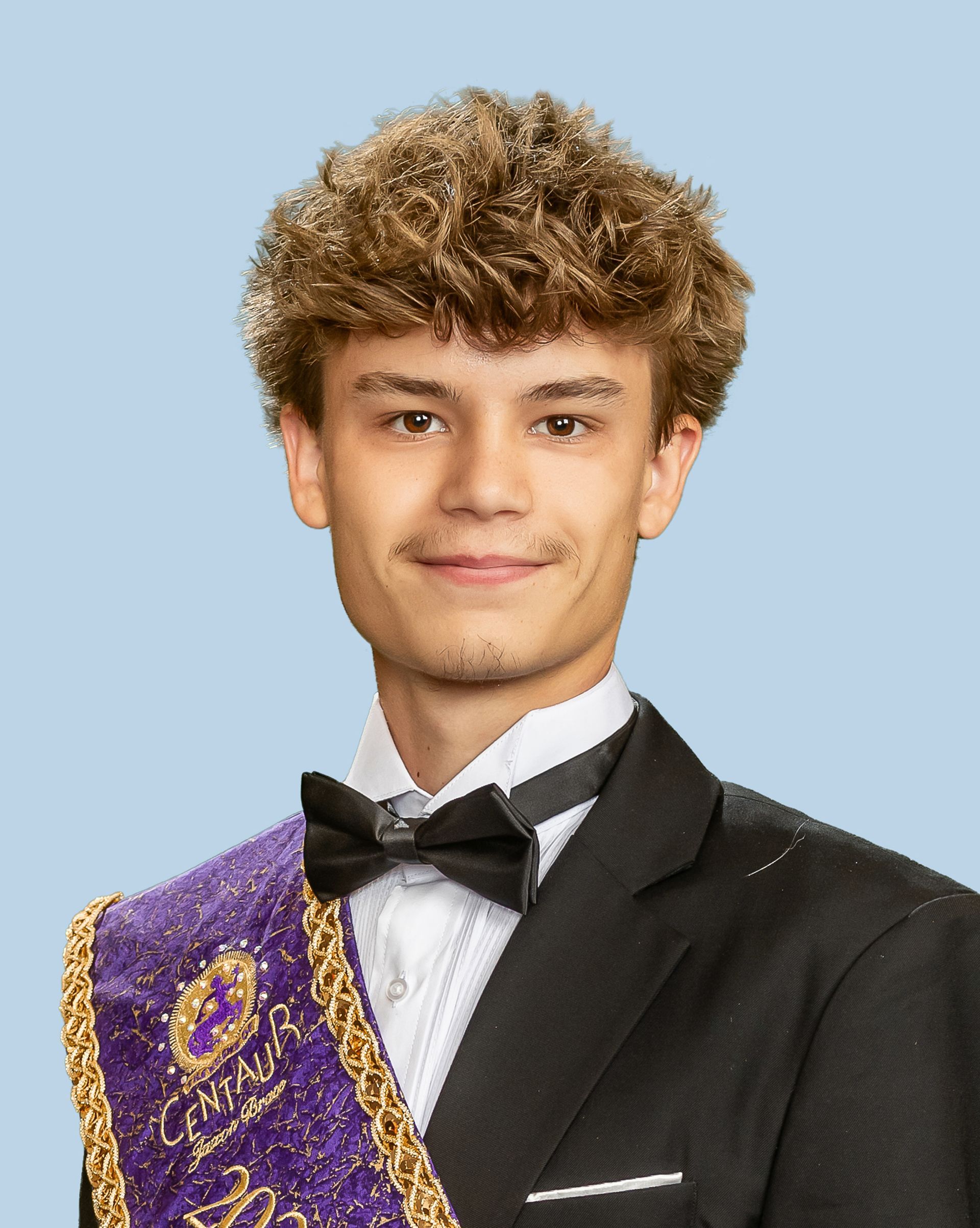 A young man in a tuxedo and bow tie is wearing a purple sash, who is a prince. 