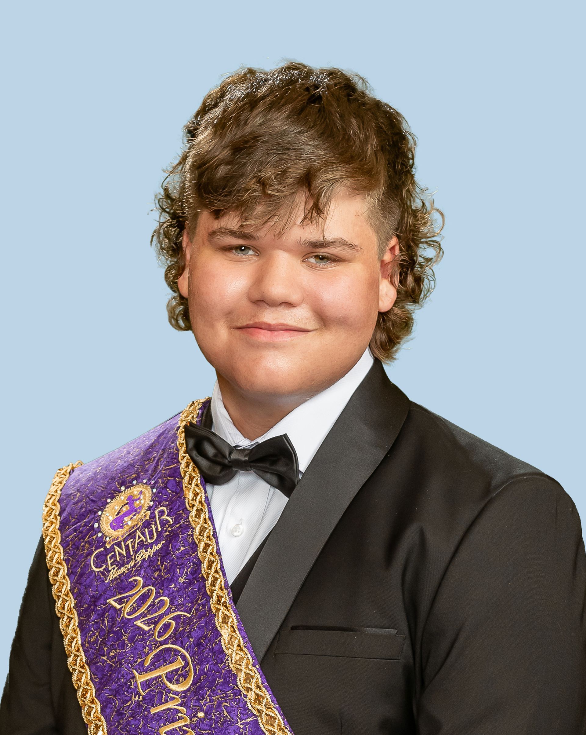 A young man in a tuxedo and bow tie is wearing a purple sash, who is a prince. 