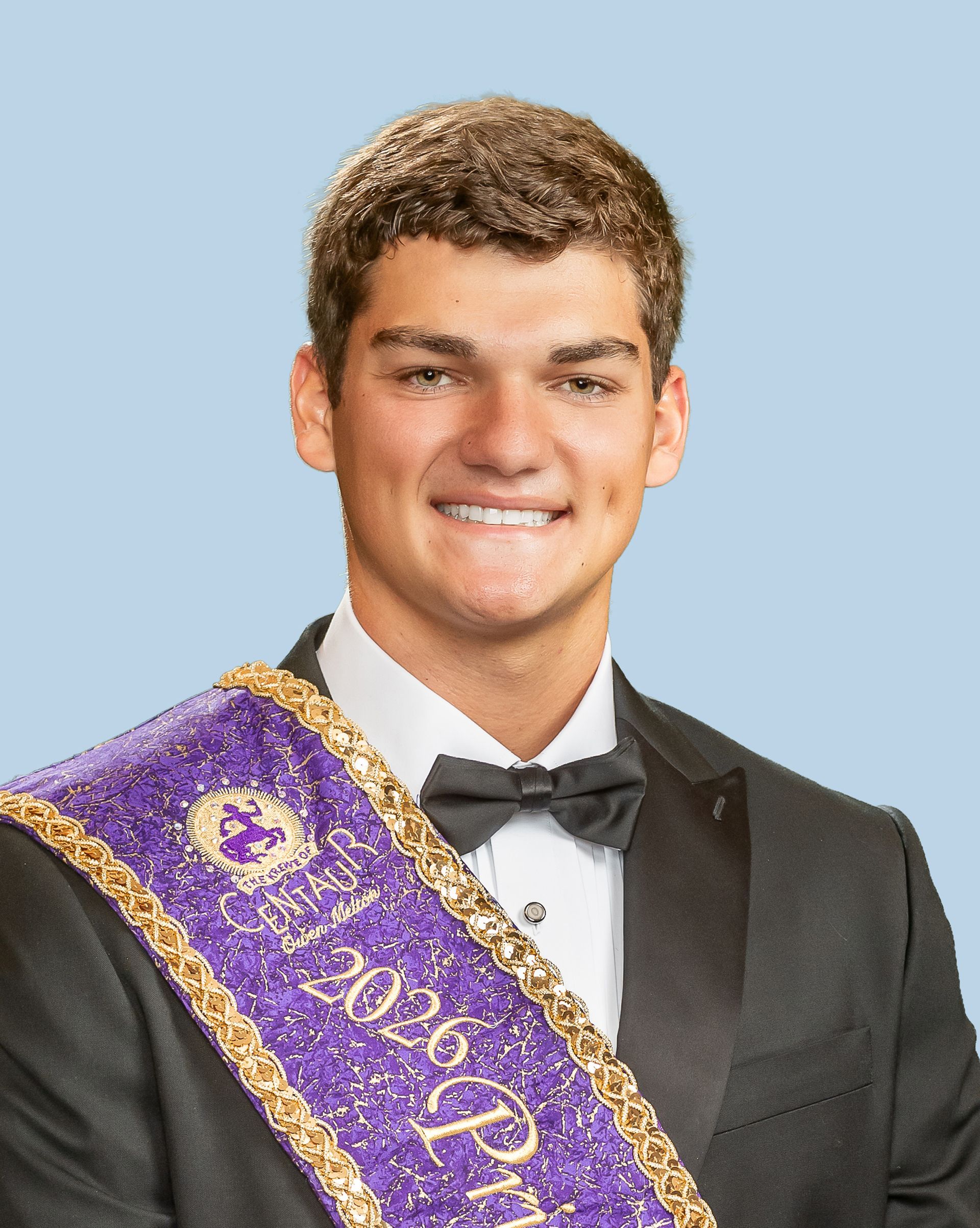 A young man in a tuxedo and bow tie is wearing a purple sash, who is a prince. 
