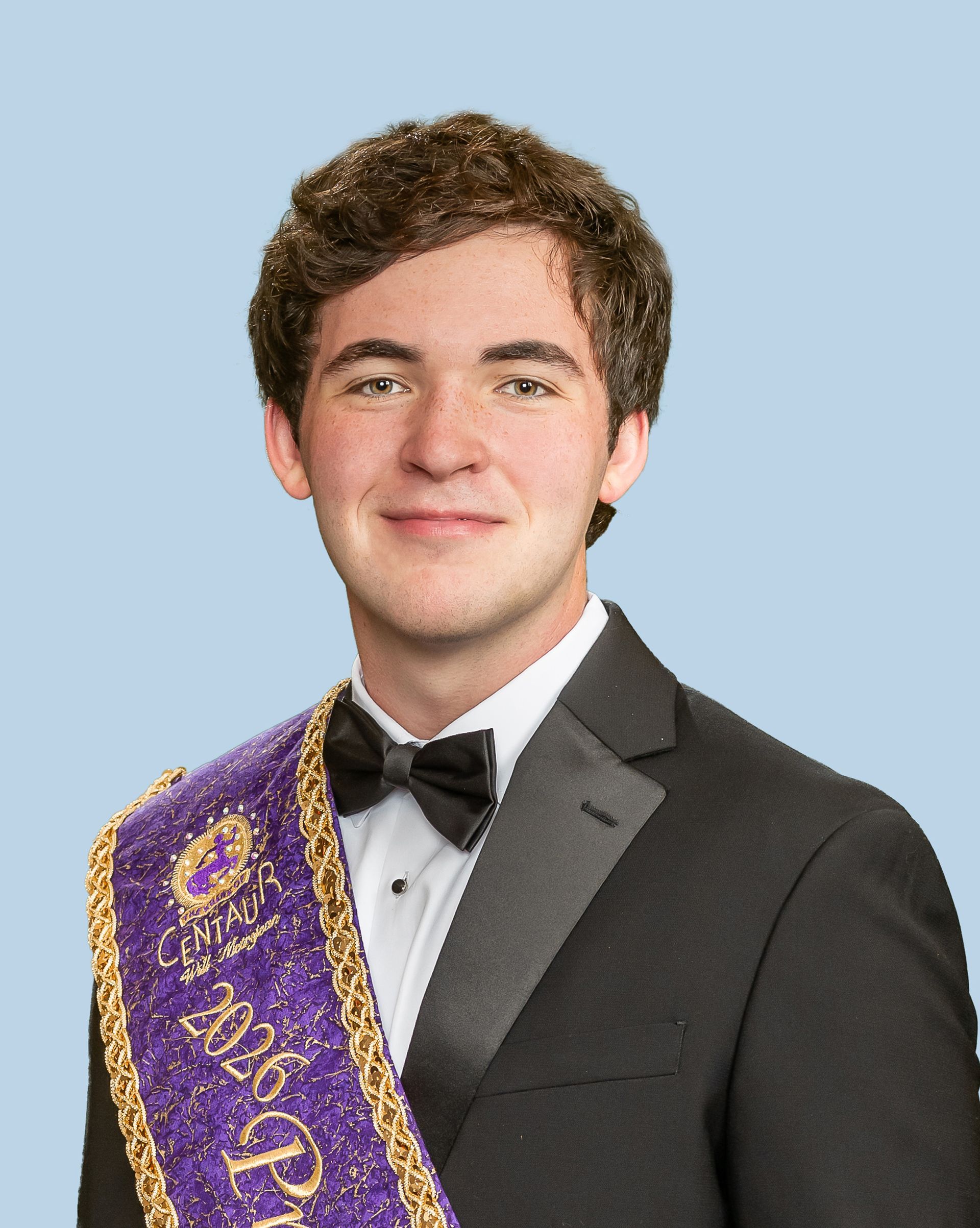 A young man in a tuxedo and bow tie is wearing a purple sash, who is a prince. 