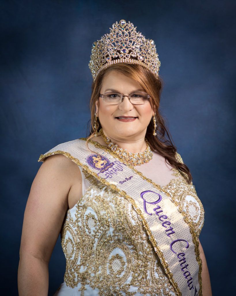 A woman wearing a crown and sash is standing in front of a blue background.