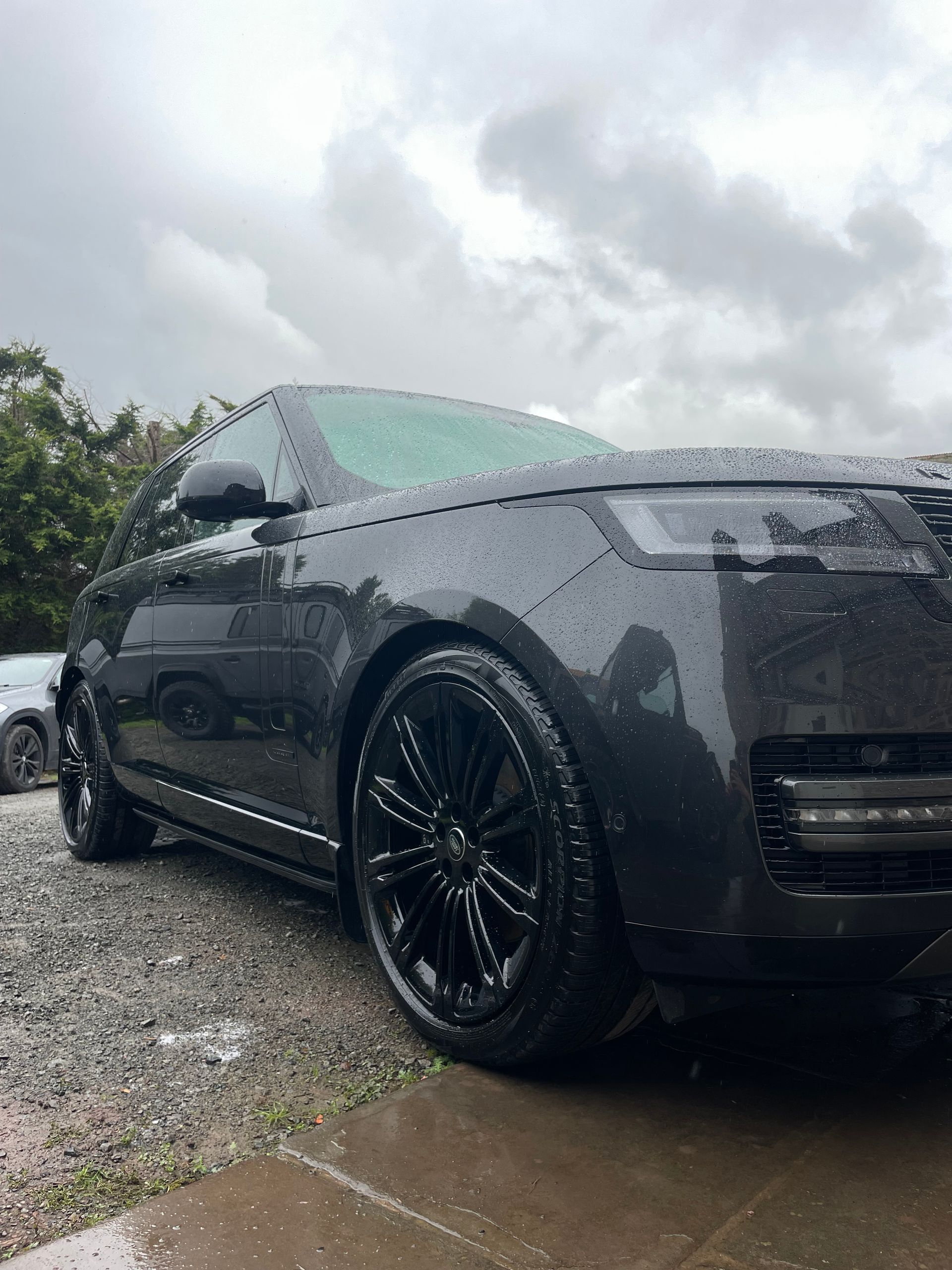 A dark gray Range Rover parked on a gravel surface under a cloudy sky, showing raindrops on the vehicle's body.