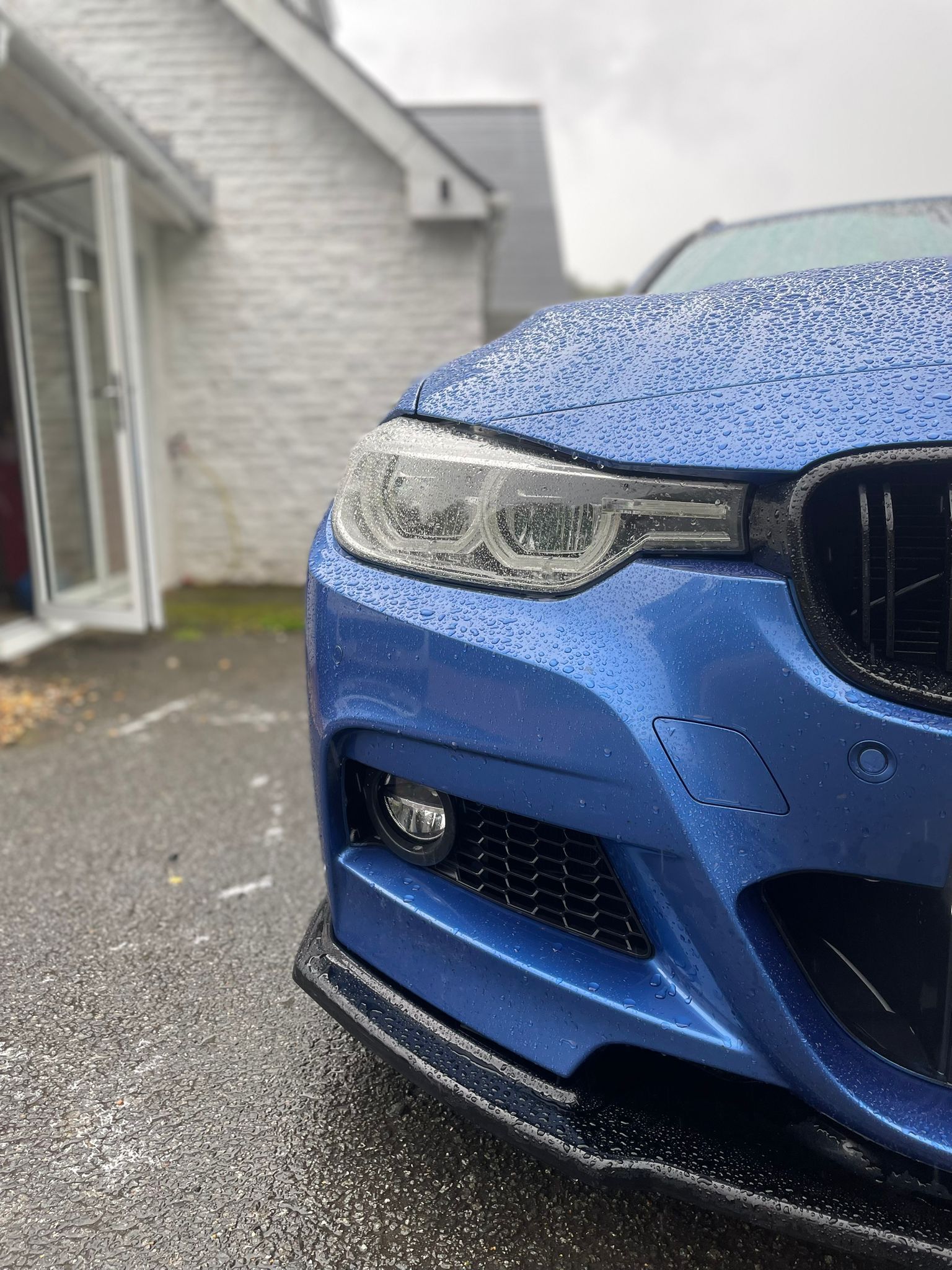 Blue BMW car front with wet surface; next to a house under overcast sky.