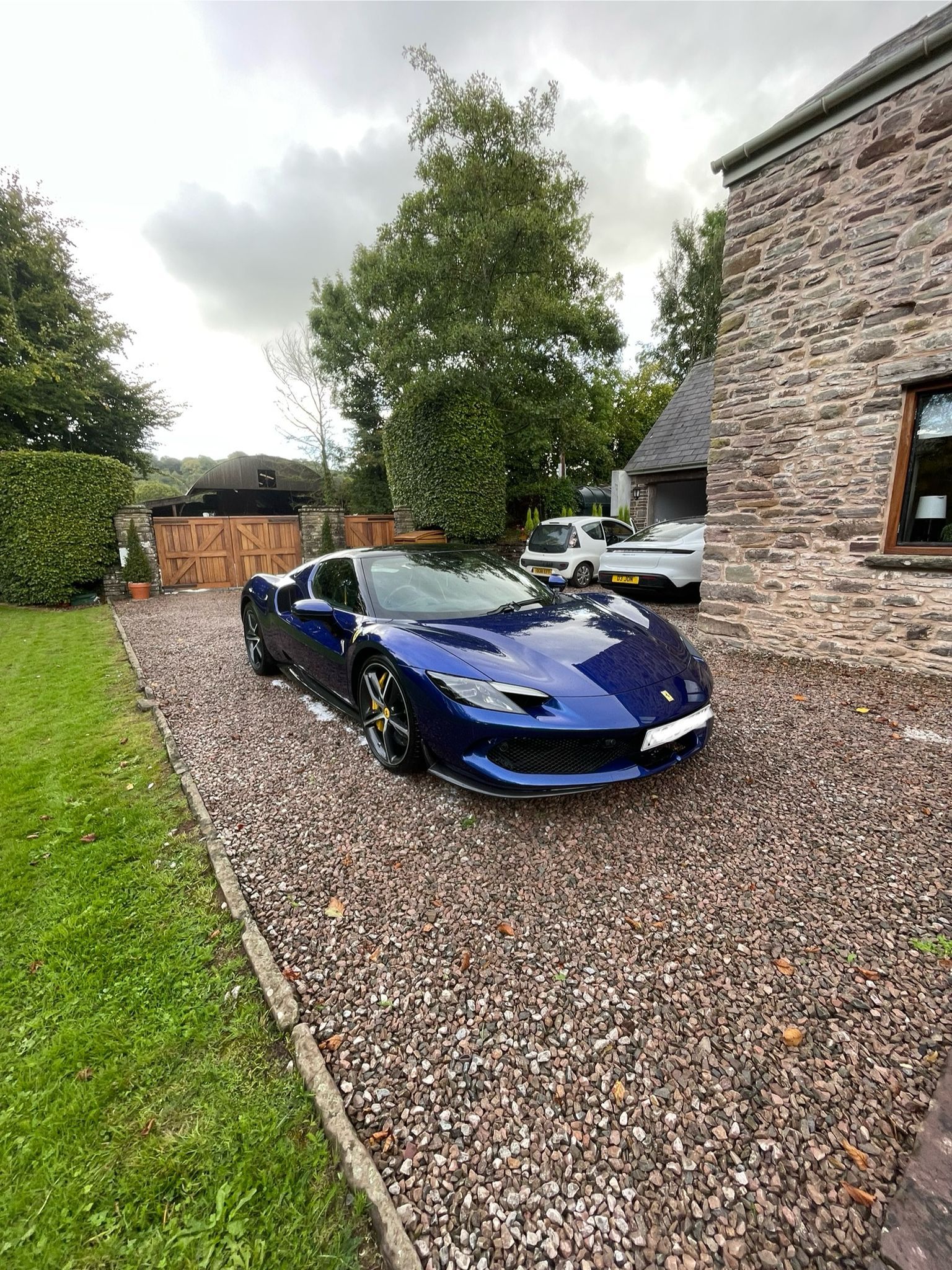 Blue sports car parked on gravel next to a stone building and lawn. Overcast sky.
