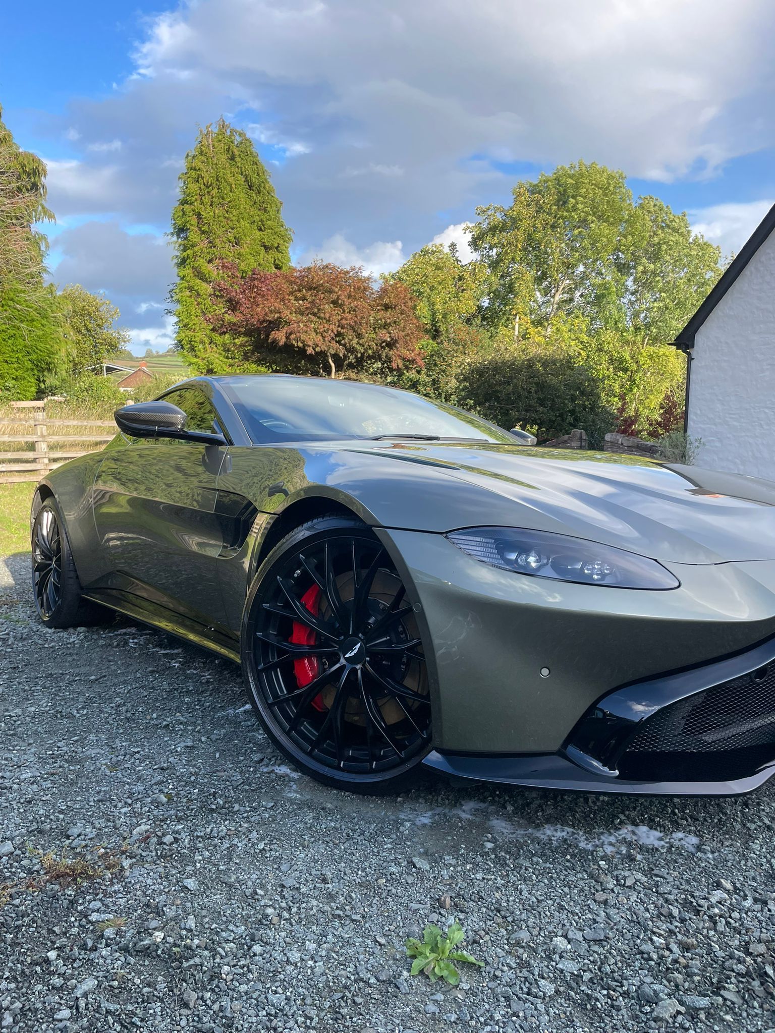 Green Aston Martin sports car parked on gravel, with black rims and red brake calipers.