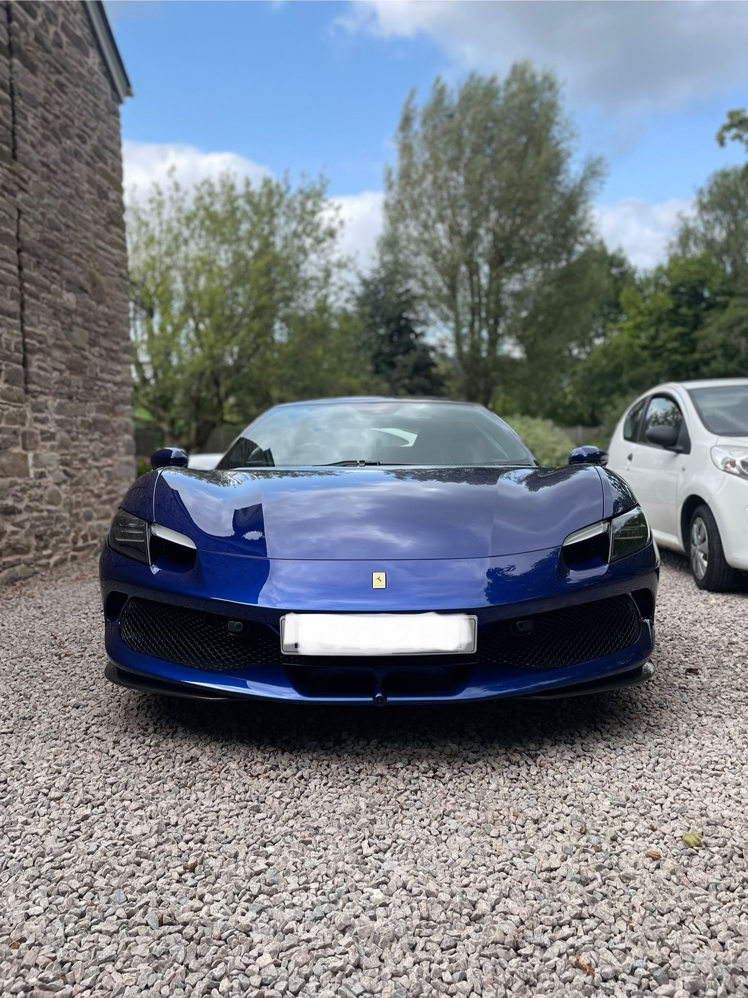 Blue sports car parked on gravel, in front of a stone building with trees in the background.