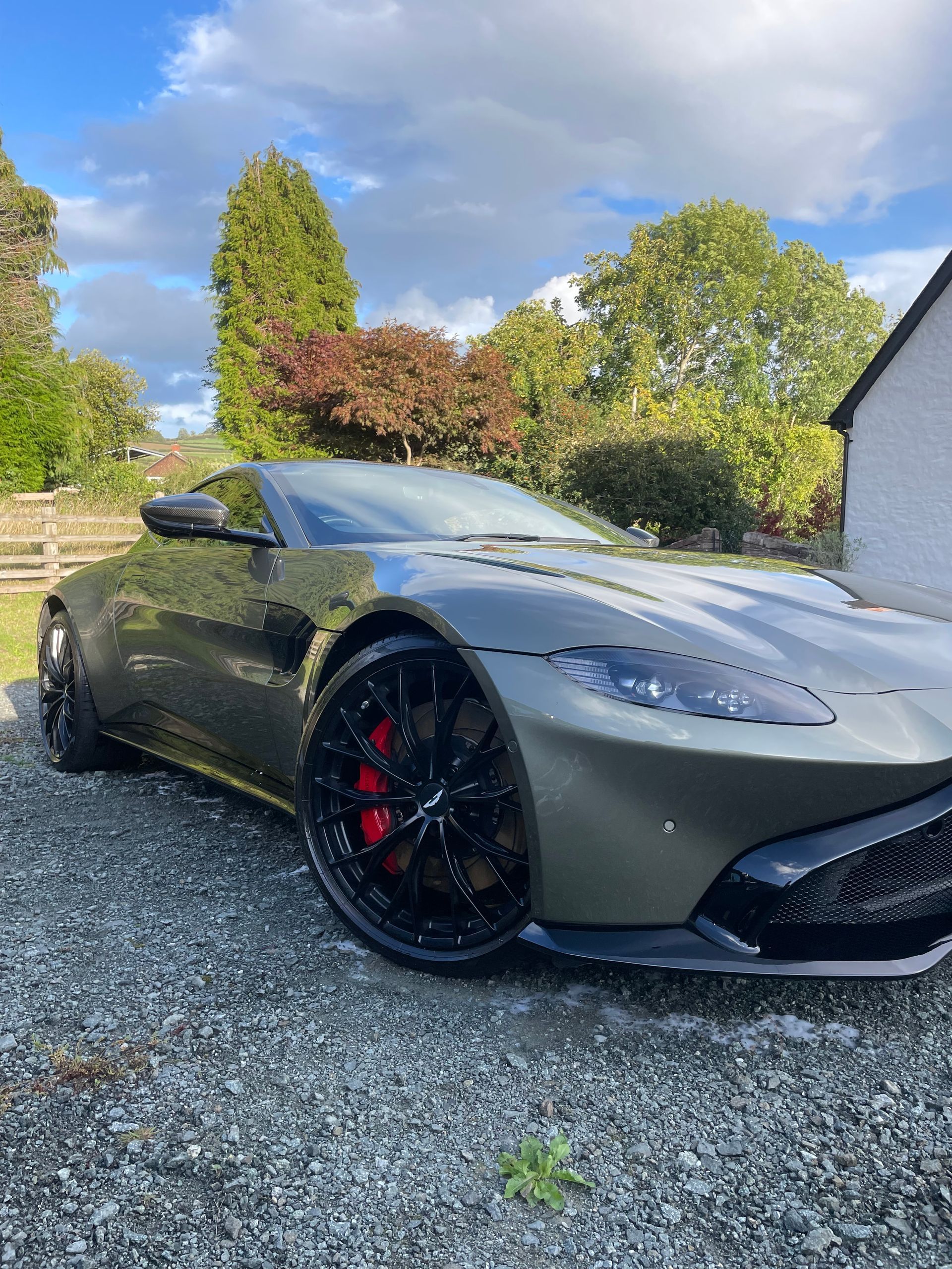 A dark grey Aston Martin sports car parked on a gravel driveway outdoors with trees and a building in the background.