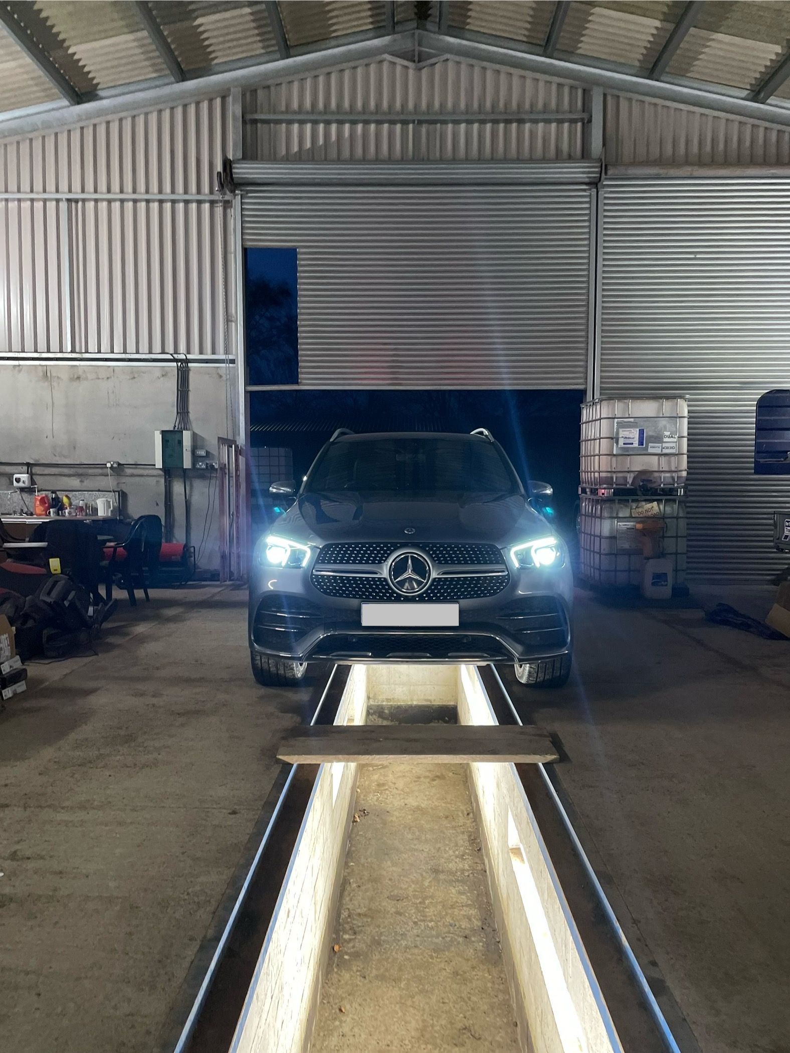 A dark Mercedes-Benz SUV parked over a lit inspection pit inside a large industrial garage.