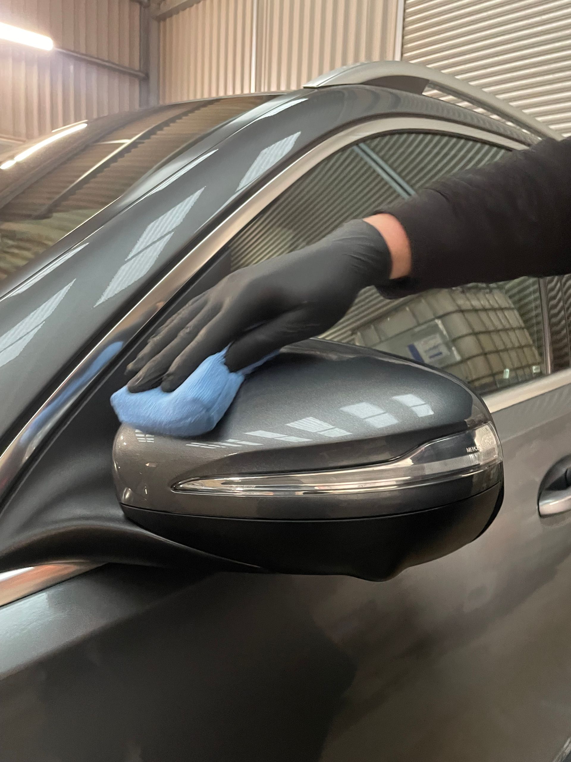 A person wearing black gloves wipes a gray car side-view mirror with a blue cloth in a garage.