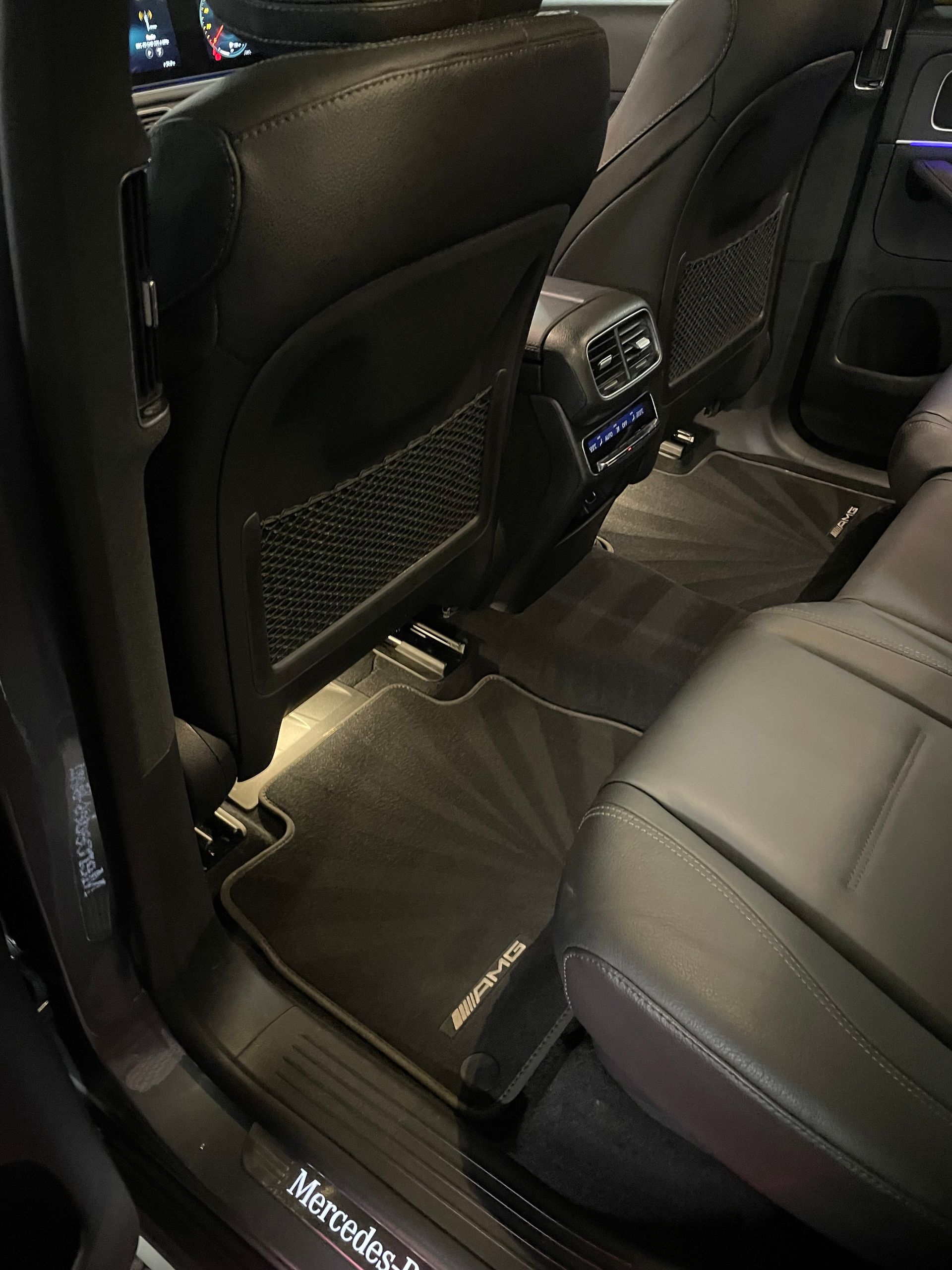 Back seat interior of a Mercedes-AMG vehicle showing black leather upholstery, branded floor mats, and climate controls.