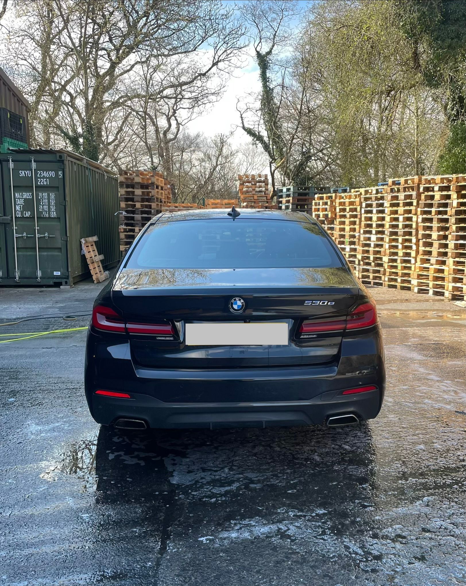 A black BMW sedan parked on a wet, gravel-covered lot in front of stacks of wooden pallets and a shipping container.
