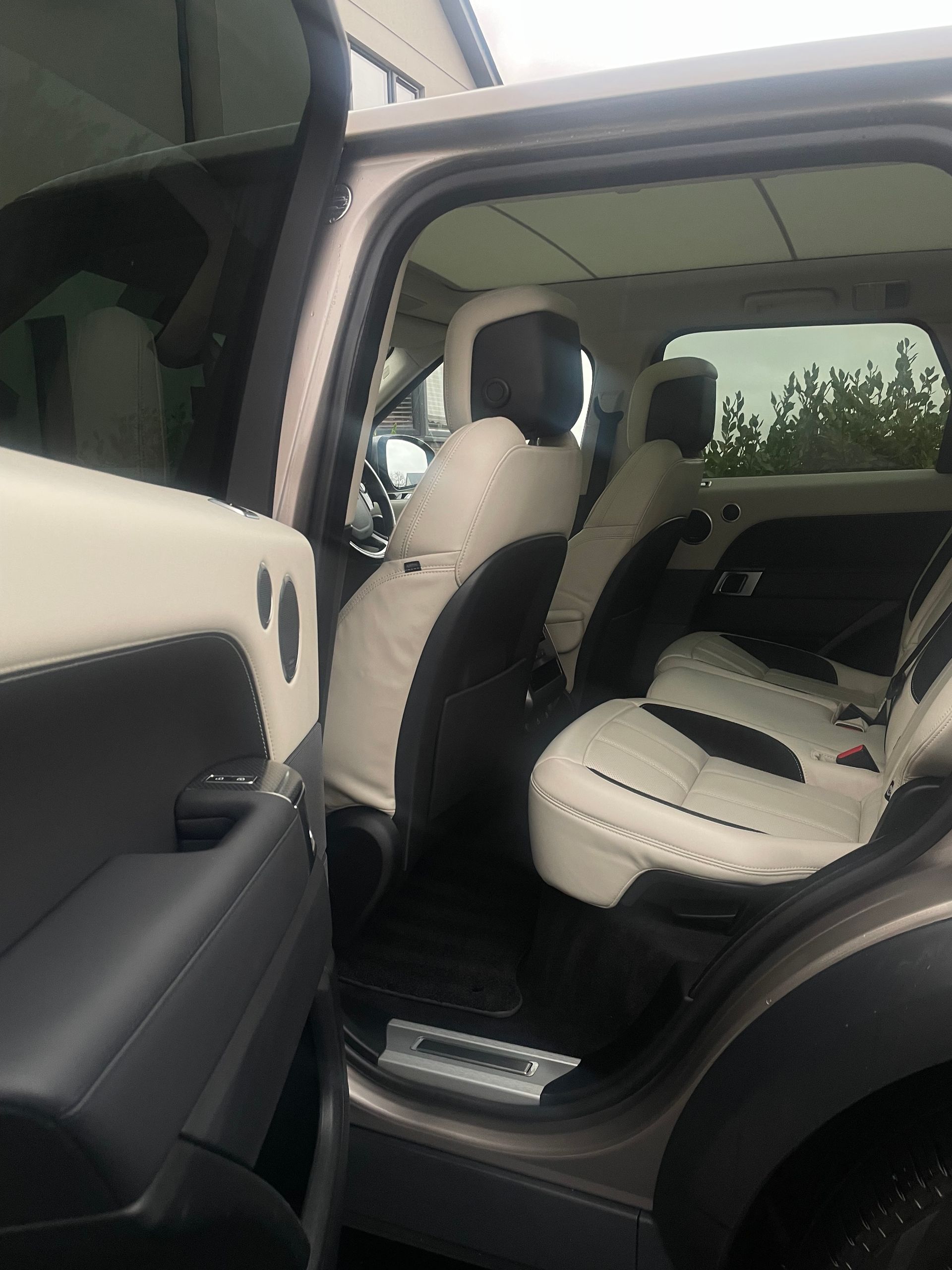 Interior view of a vehicle through an open rear door, showing cream and black leather seats and a panoramic glass roof.