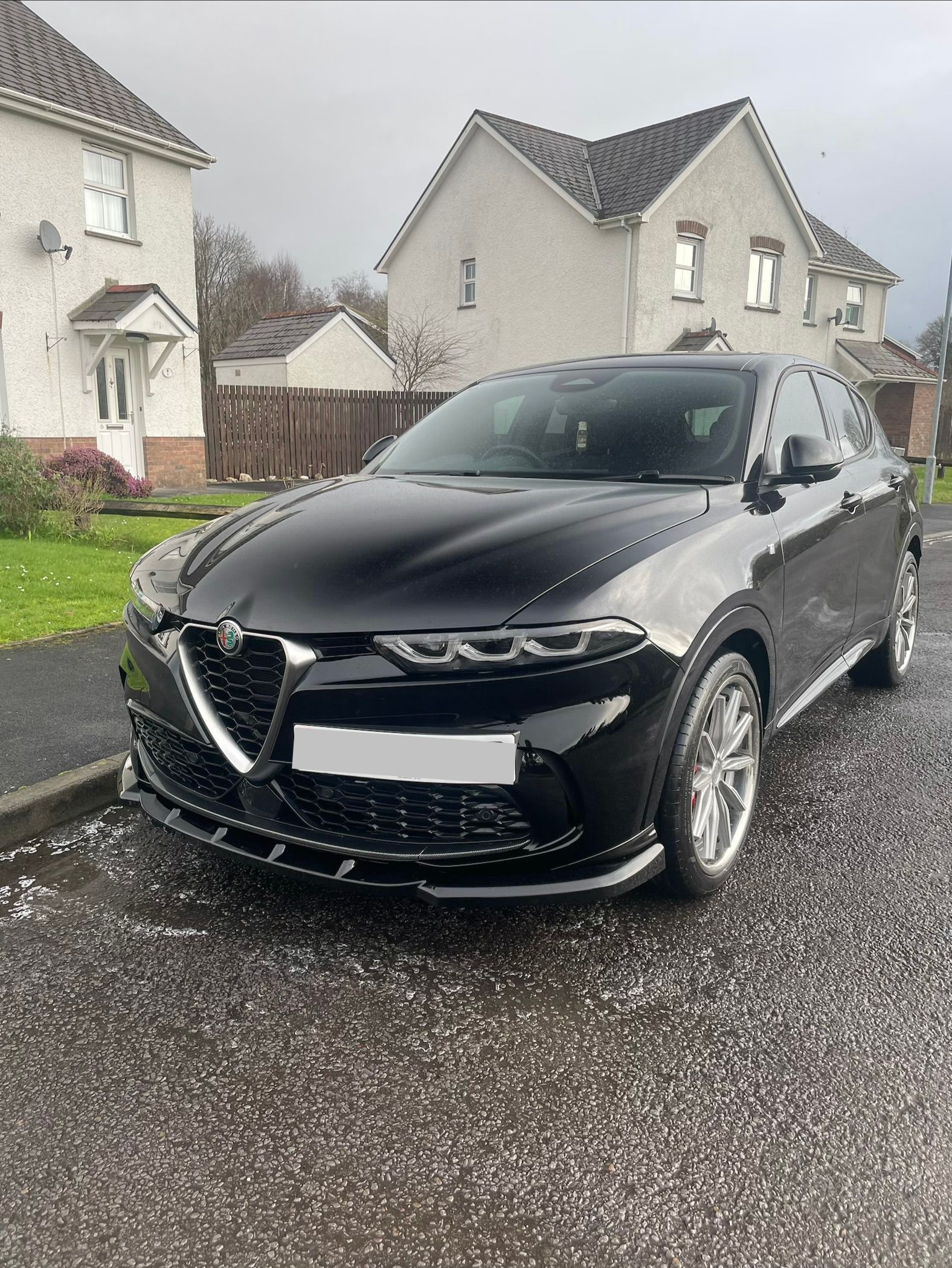 A black Alfa Romeo Tonale parked on a paved driveway in front of suburban houses.