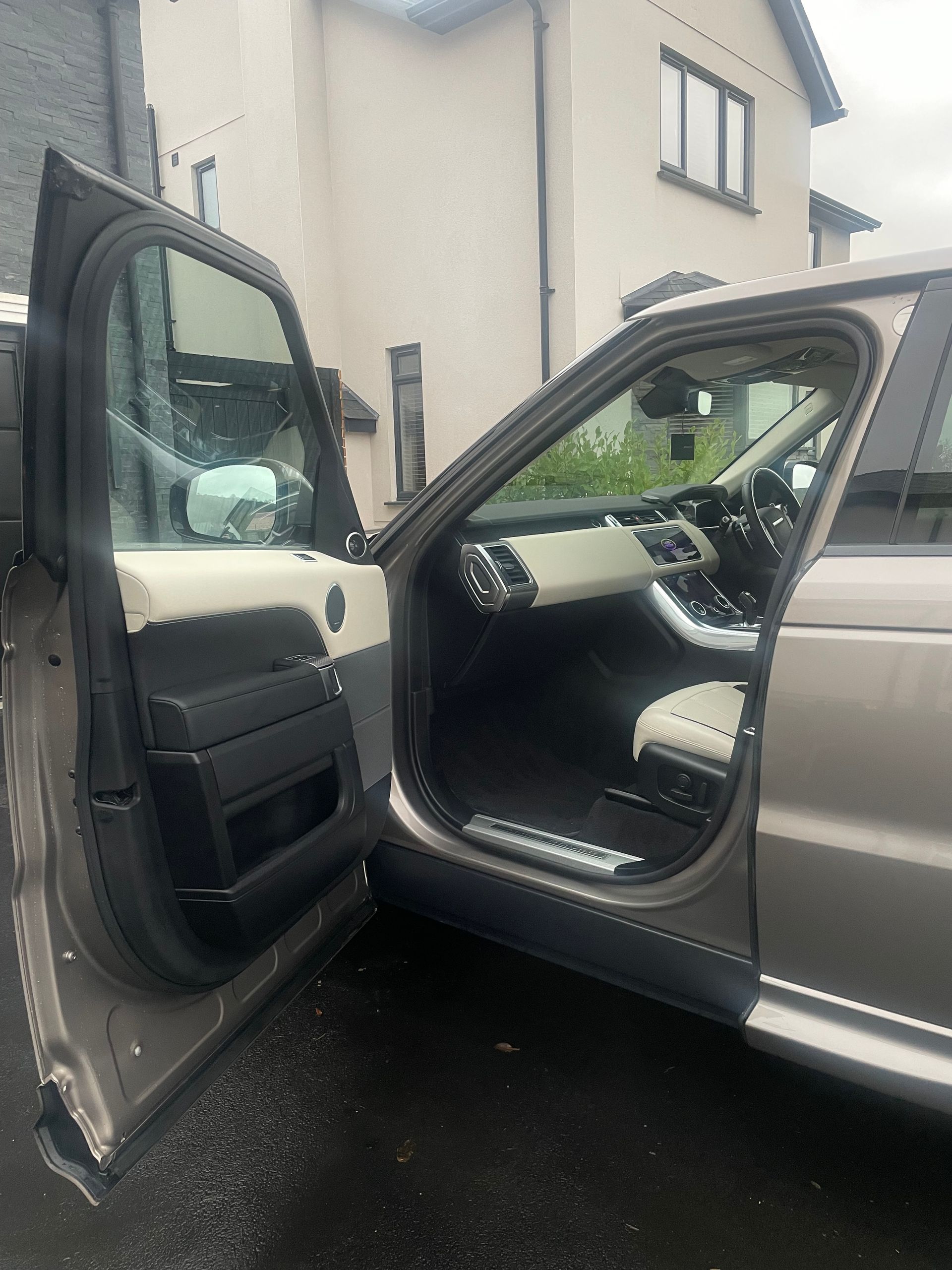 An open front door of a bronze Land Rover Range Rover, showing a black and beige interior parked in a residential driveway.