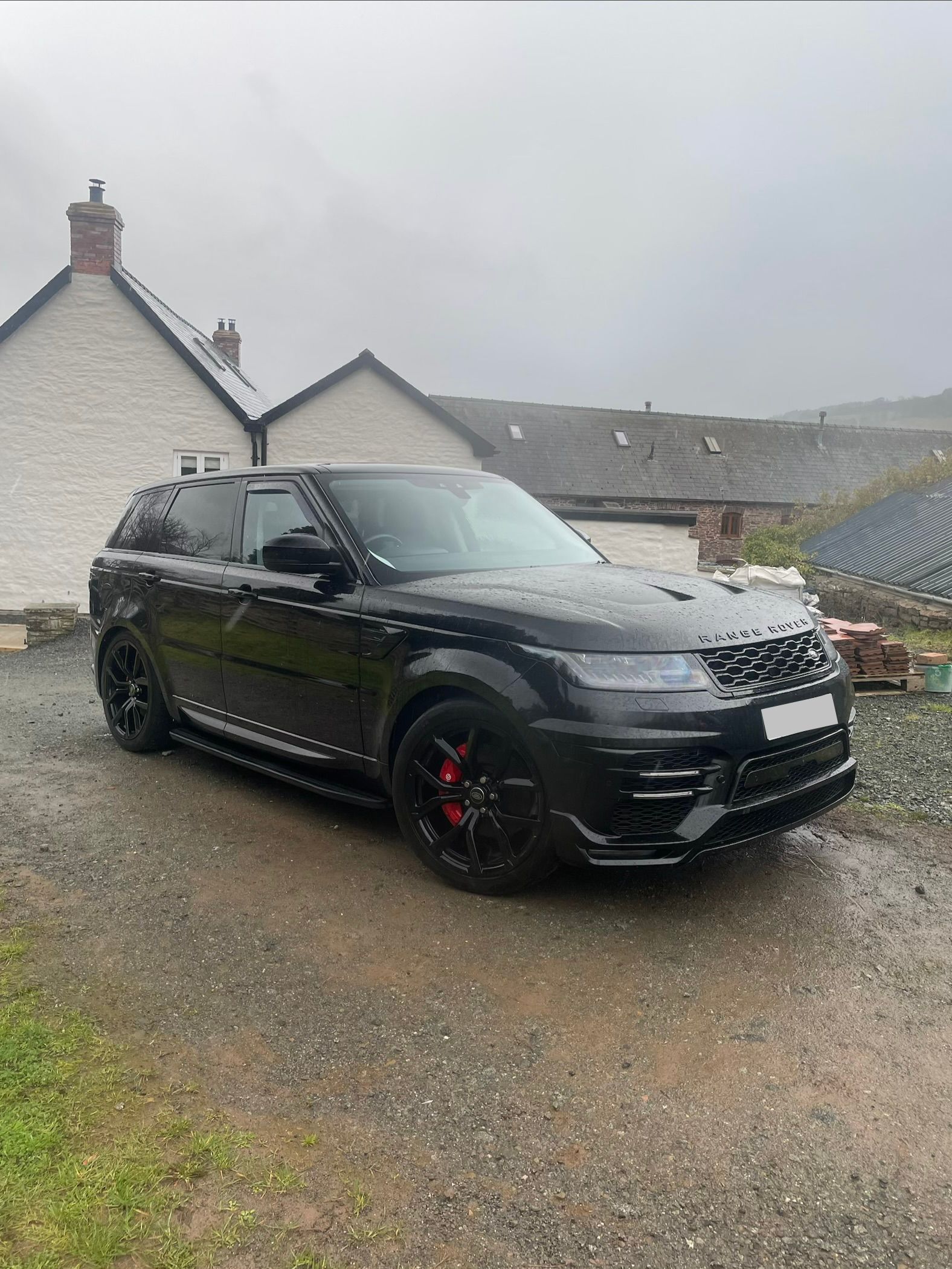 A black Range Rover parked on a wet, gravel driveway in front of a stone cottage on a cloudy day.