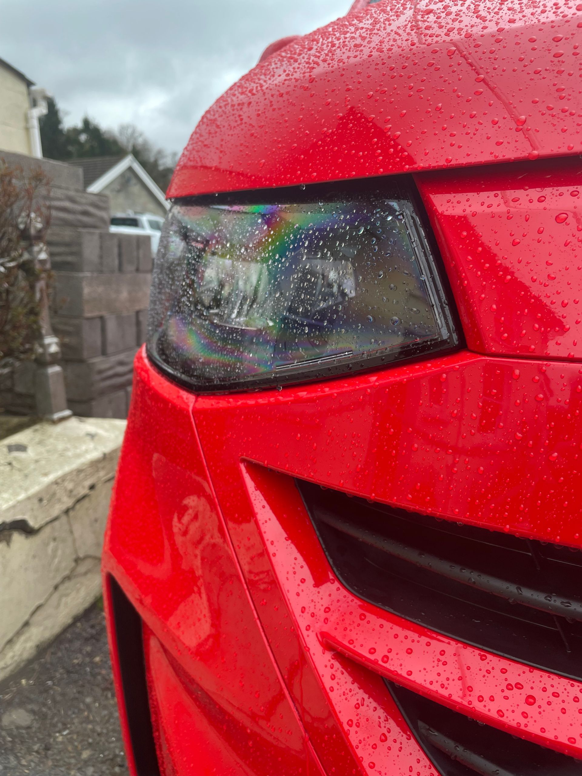 Close-up of a bright red car headlight covered in raindrops, parked outdoors on an overcast day.