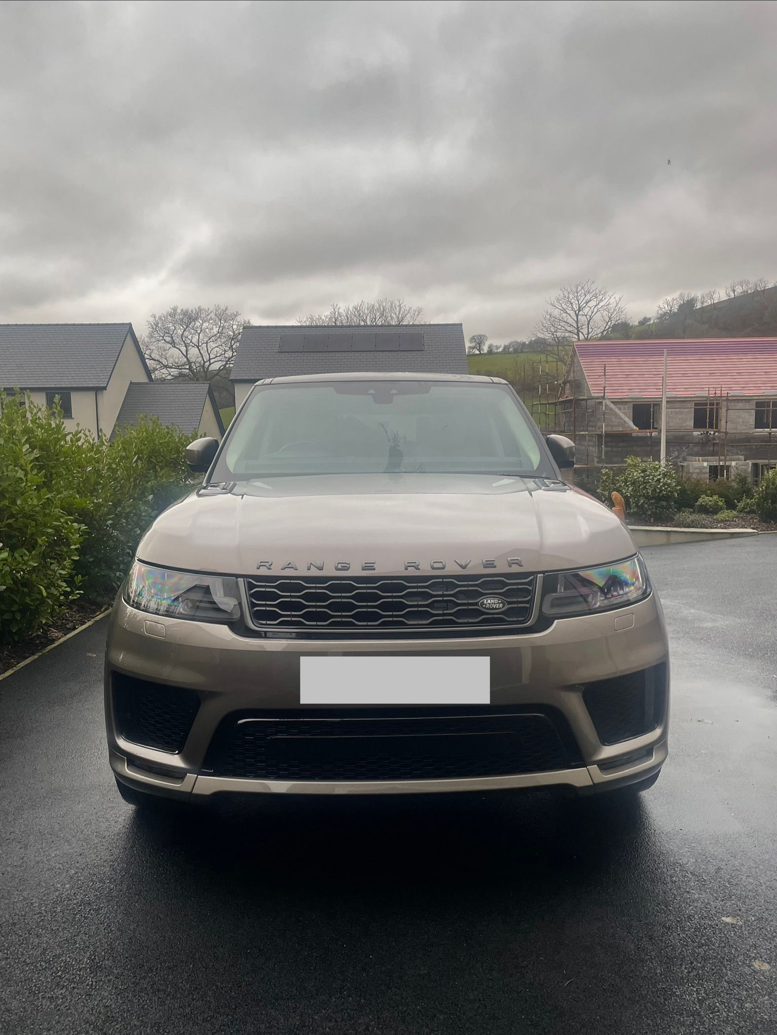 A taupe-colored Range Rover parked on a wet asphalt driveway in front of houses under a cloudy sky.