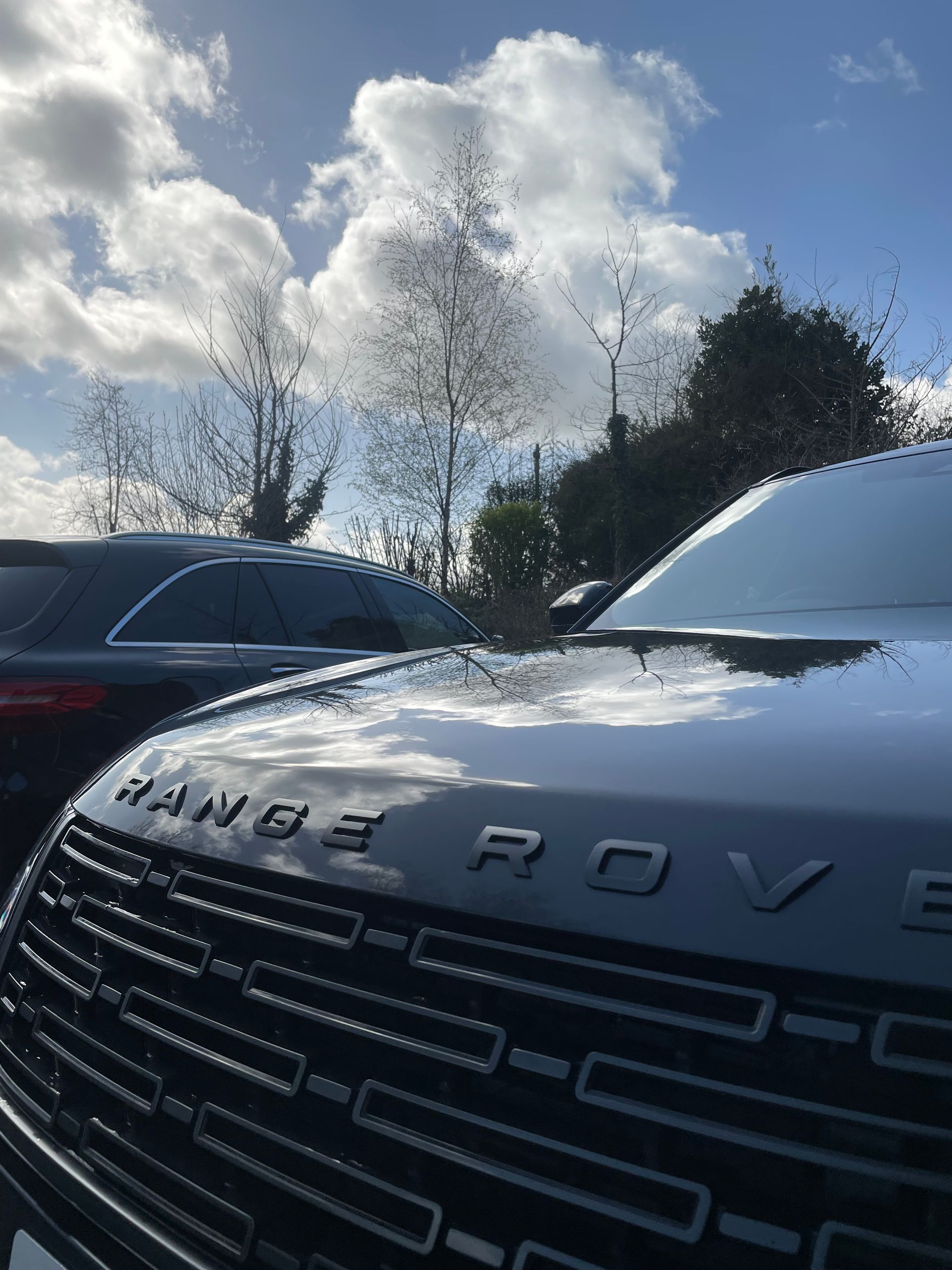 Close-up of a black Range Rover hood and grill, reflecting a sunny blue sky with clouds, parked near trees.
