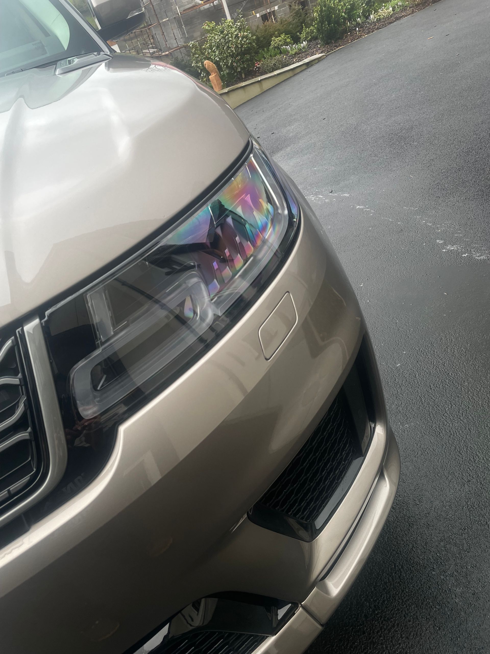 Close-up of a tan Range Rover’s front headlight and bumper on a paved driveway.