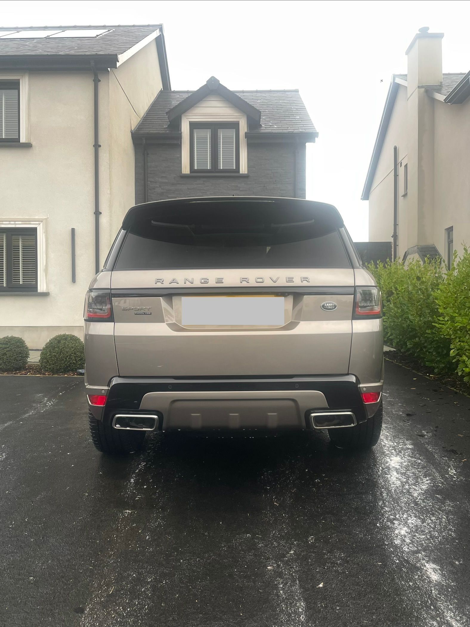 A gold Range Rover parked in a driveway between two residential houses on a gloomy day.