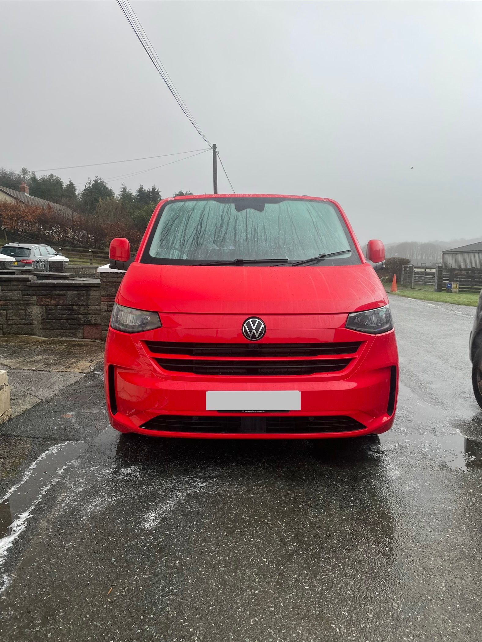 A bright red Volkswagen Transporter van parked on a wet, paved surface outdoors during a rainy or overcast day.