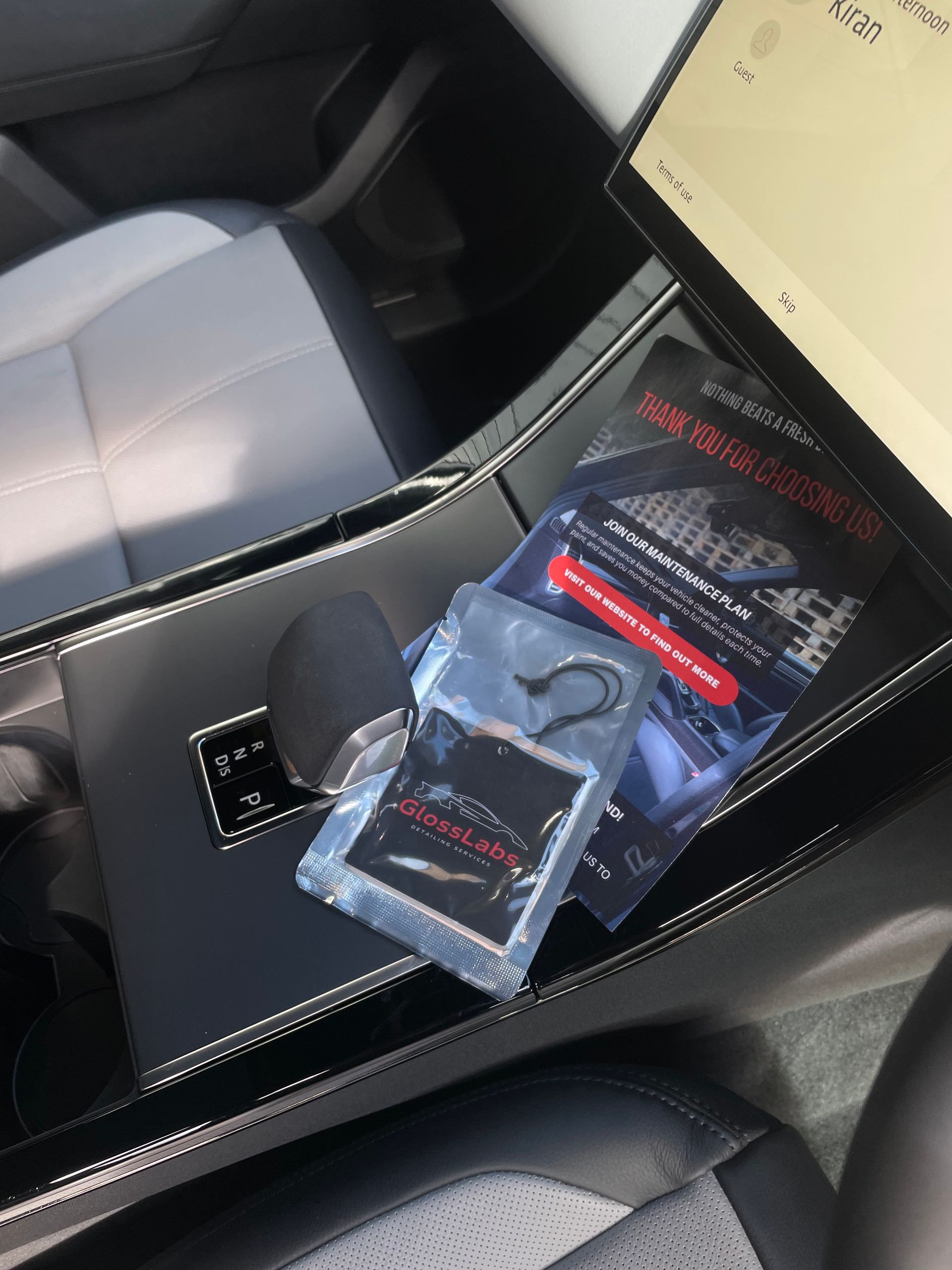 A car interior center console with a gear shifter, a packaged air freshener, and a promotional pamphlet.
