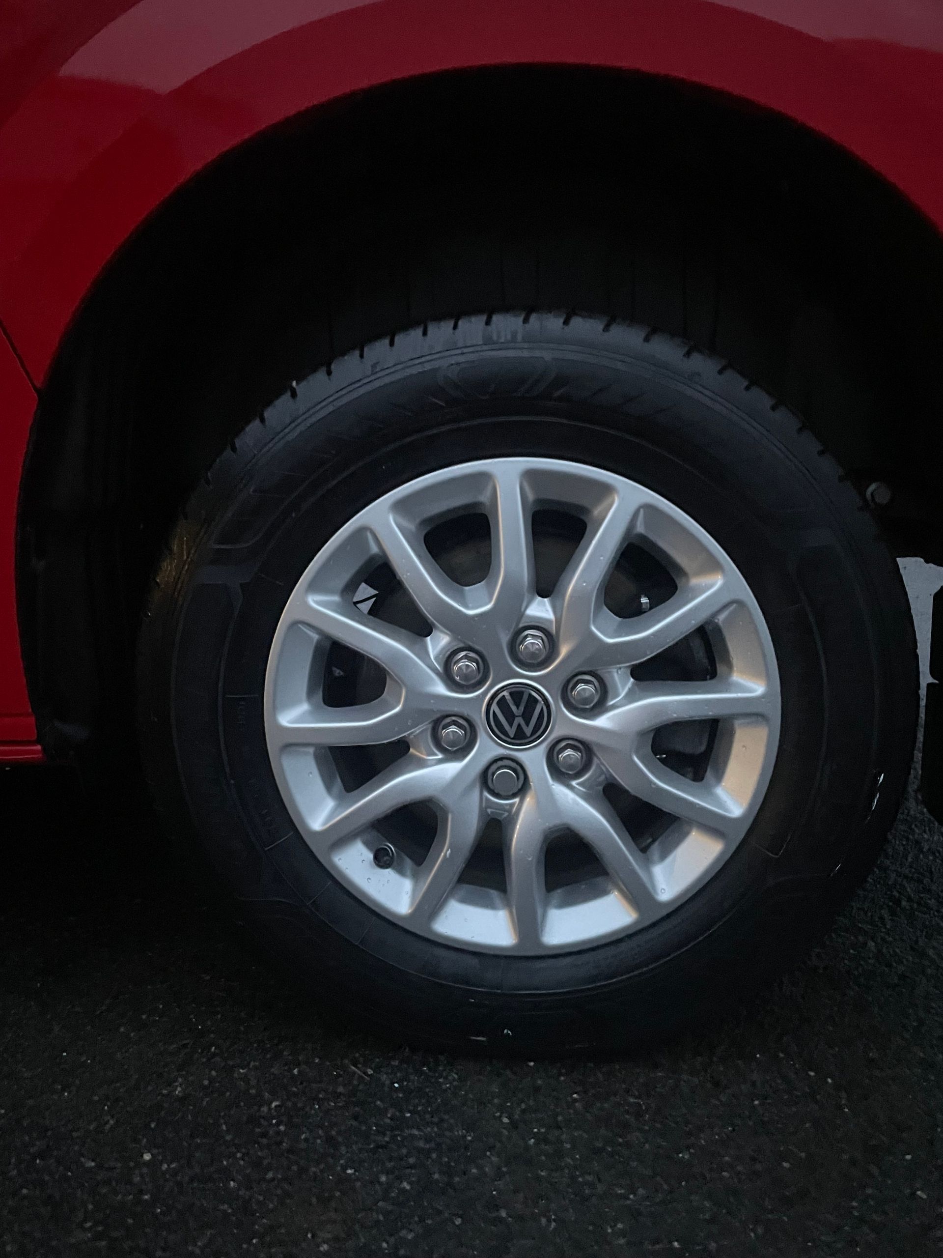 A Volkswagen car tire and silver multi-spoke wheel against a red vehicle fender on asphalt.