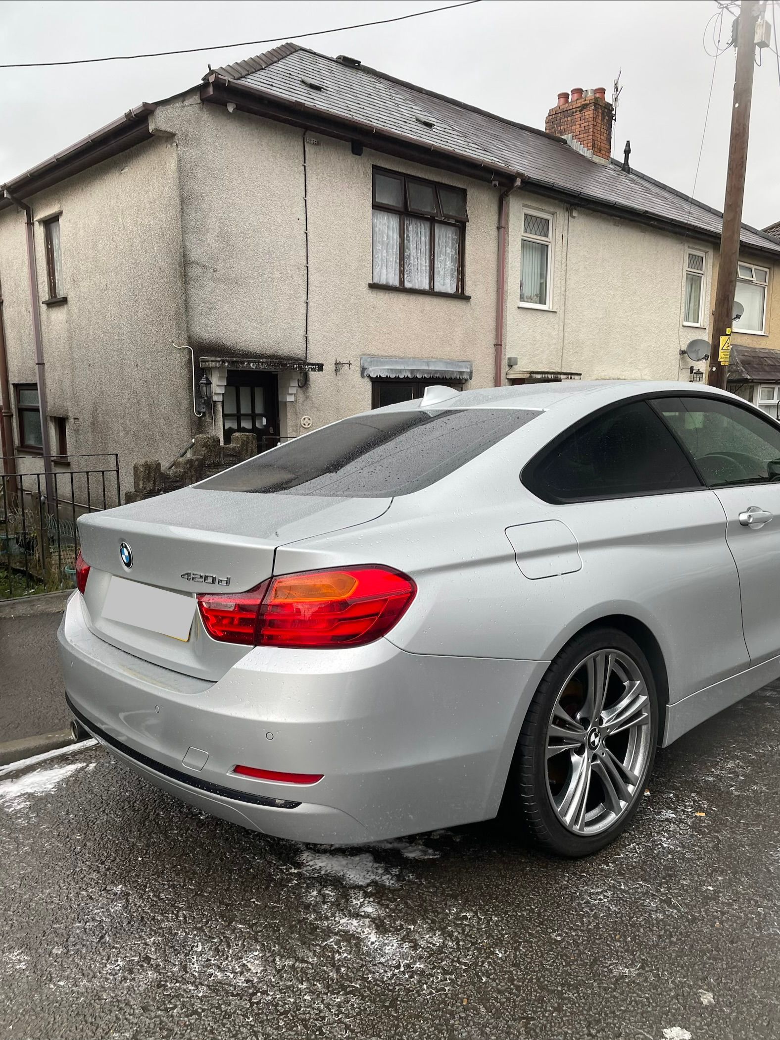 A silver BMW coupe parked on a residential street in front of a white two-story house on a gloomy, overcast day.