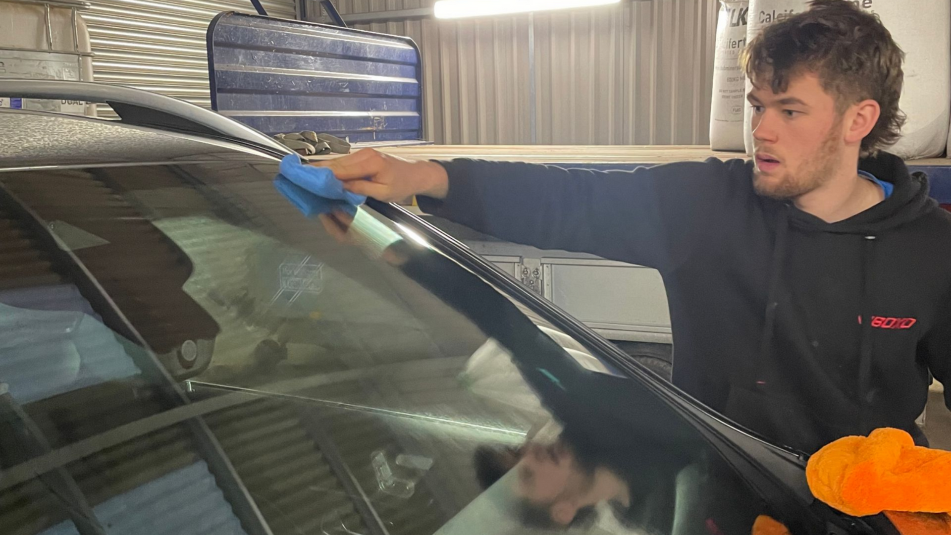 Man cleaning a car windshield with a blue cloth in a garage.