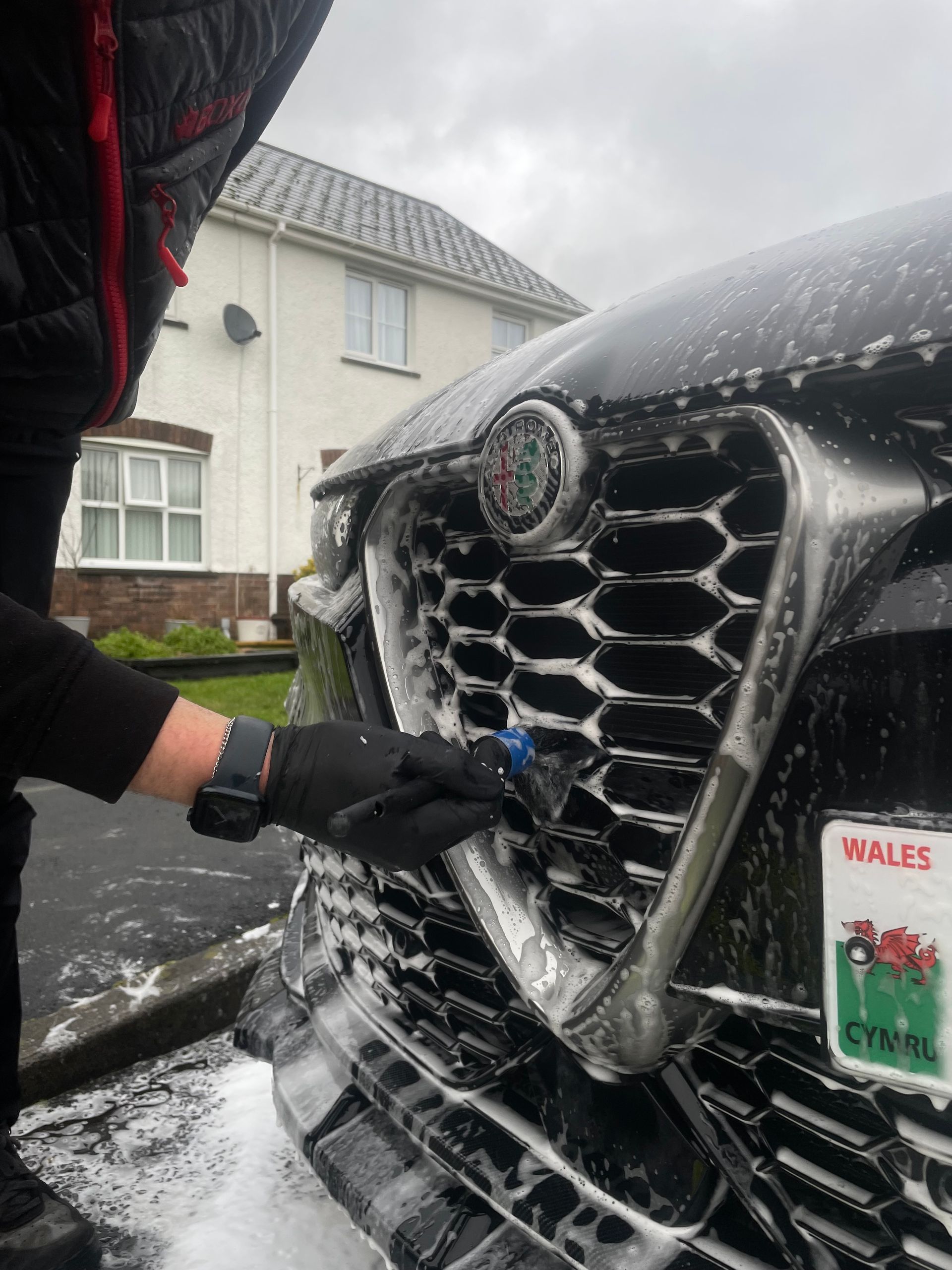 A person in a black glove uses a detailing brush to clean the foam-covered grille of a black Alfa Romeo car outdoors.