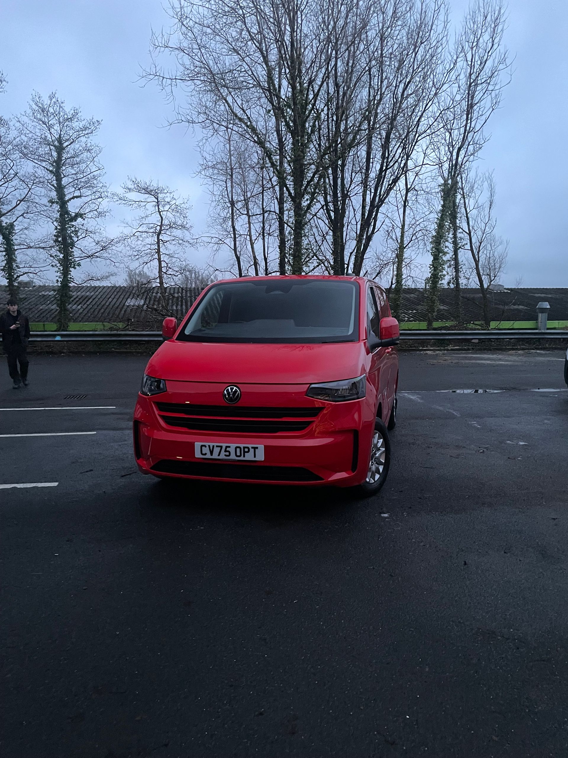 A bright red van parked in an asphalt parking lot during the day, with leafless trees and a fence in the background.