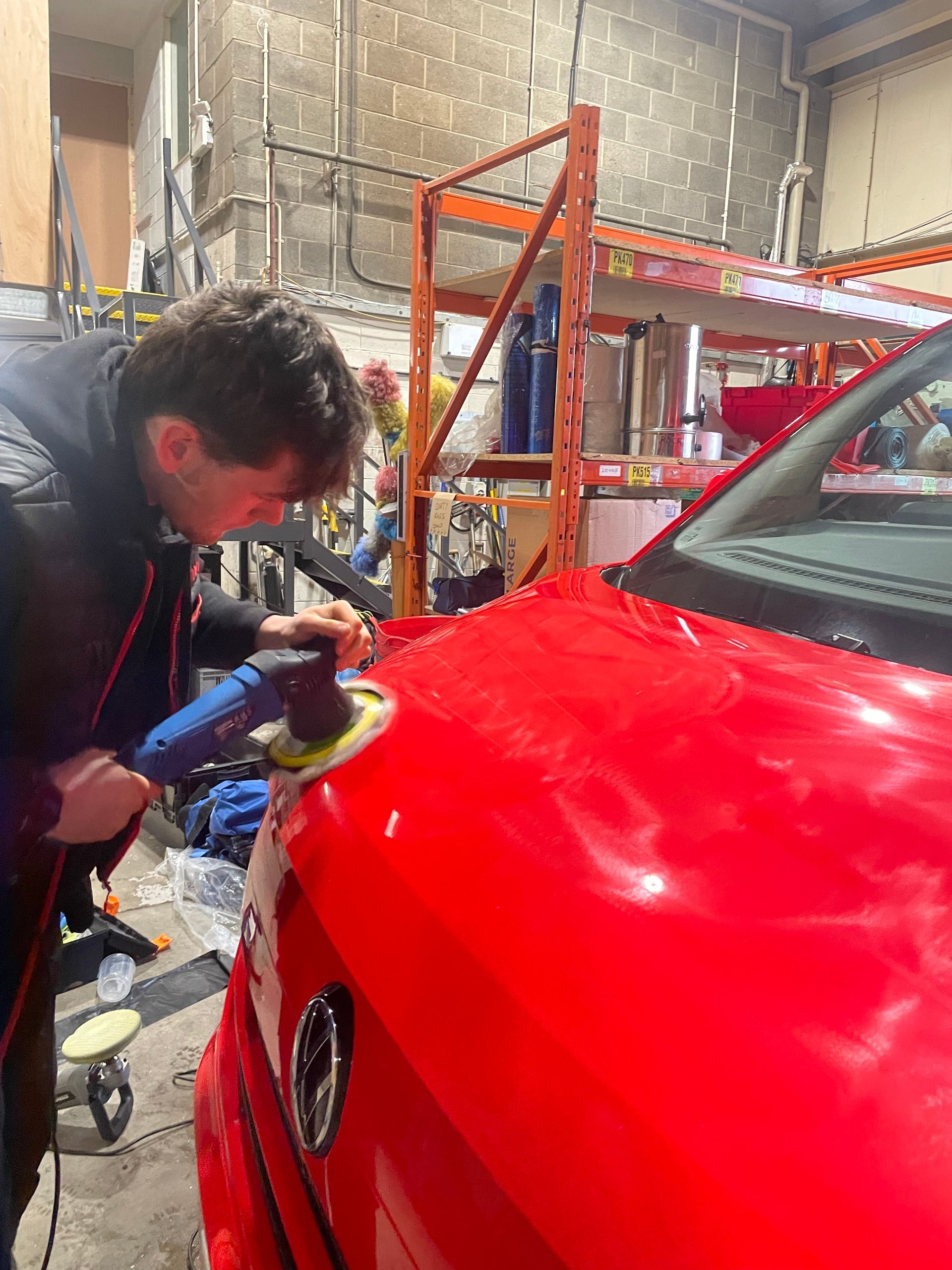 A person using a power buffer to polish the hood of a red car inside a workshop.