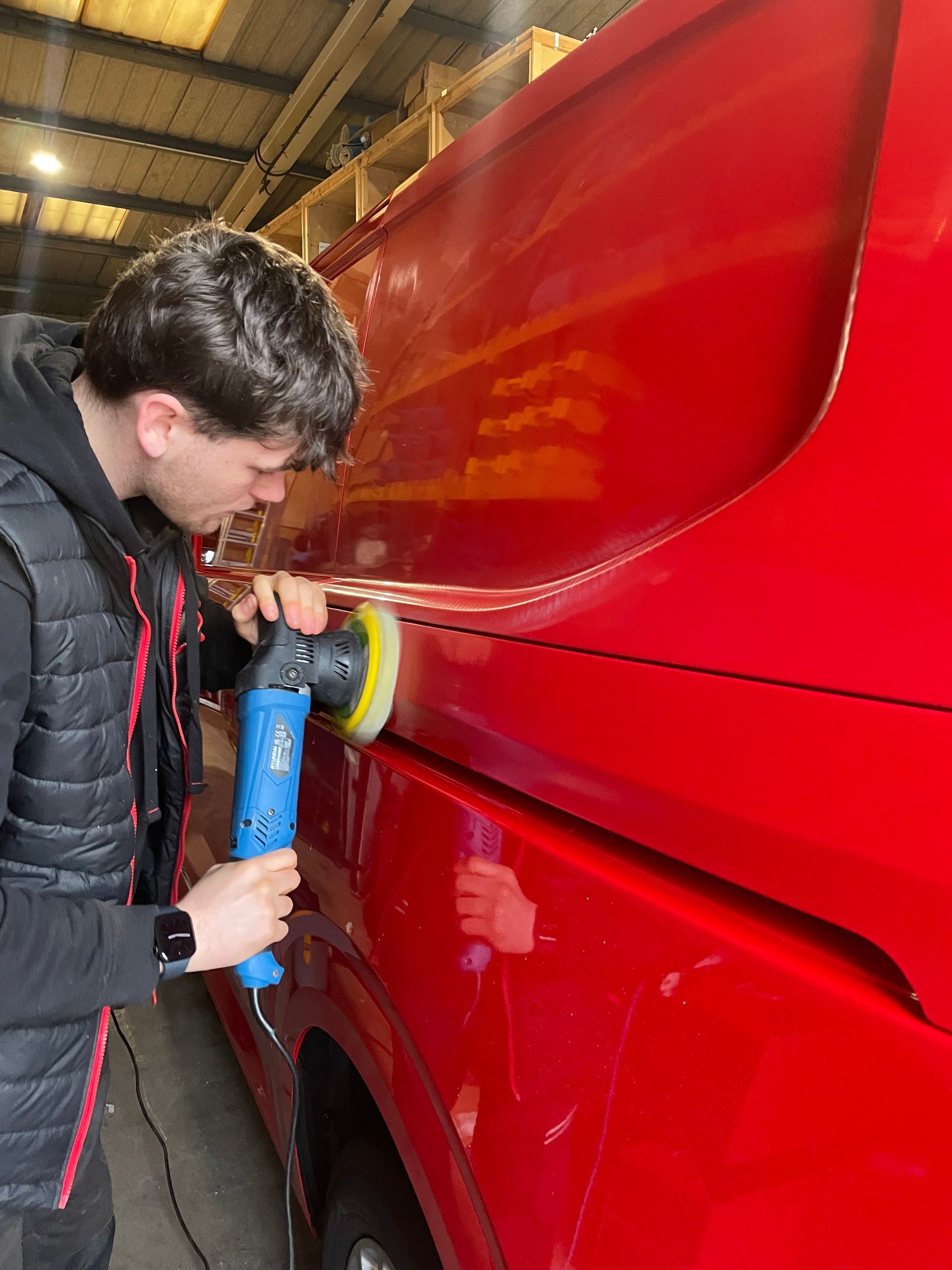 A person using a blue power polisher on the red side panel of a vehicle in an indoor garage.